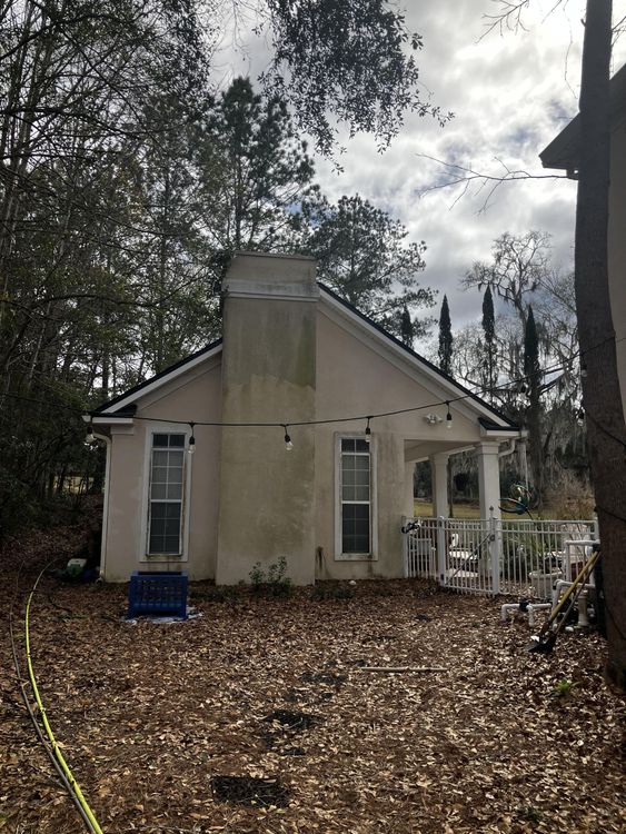 A white house with a chimney and a porch is surrounded by trees and leaves.