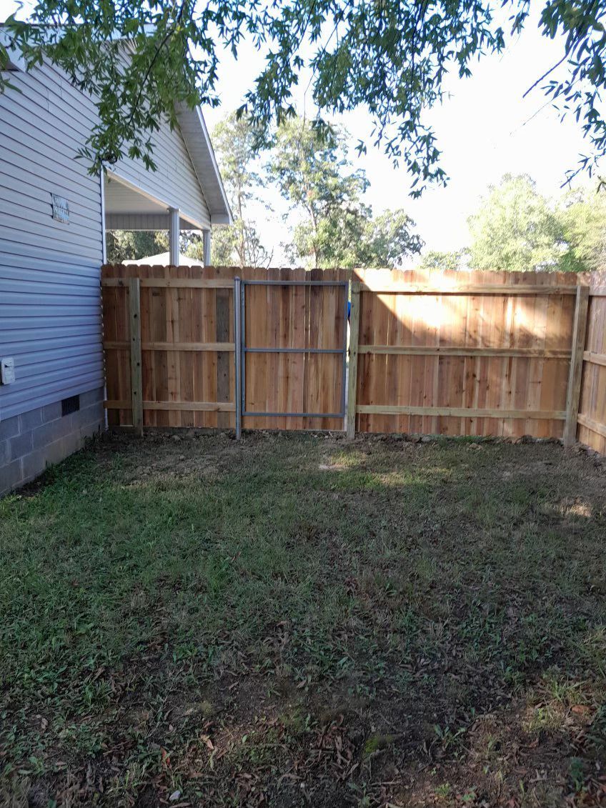 Backyard with brown wooden fence and gate. Green grass. Light blue siding of a house on the left.
