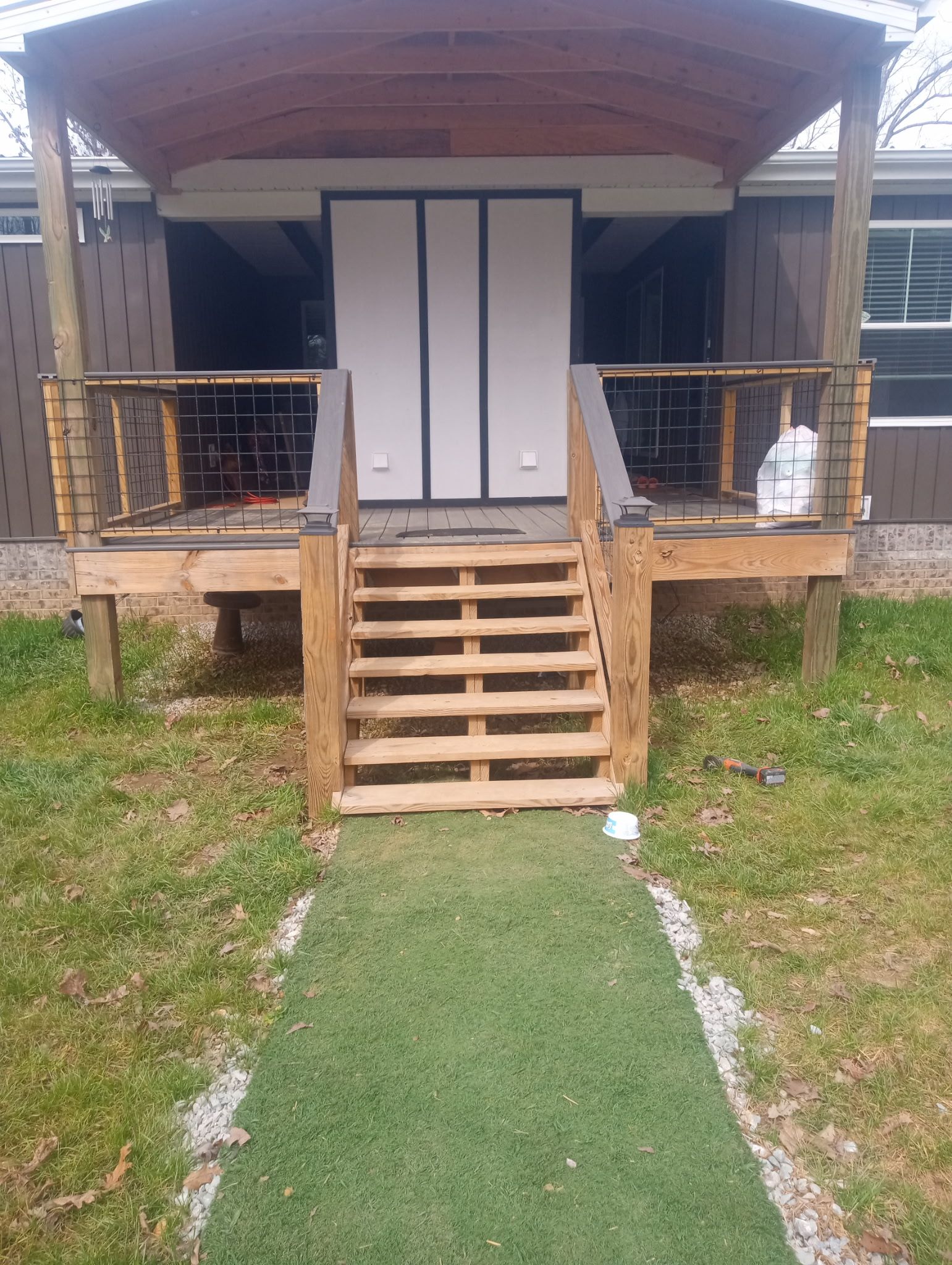 Wooden porch with steps leading to a house. Green grass pathway. Brown house with white doors.