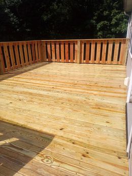 Wooden deck with vertical railing. Sunlit, surrounded by trees.