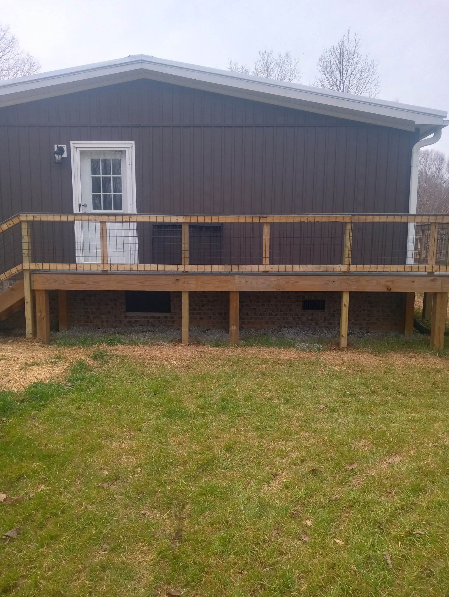 Brown house with wooden deck, wire railing, and white door, set in a grassy yard.