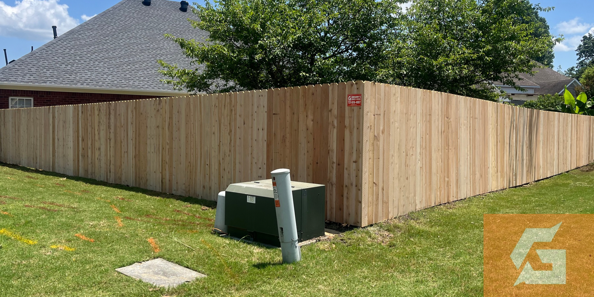 Wooden fence surrounding a yard, with a gray roof in the background and greenery on a sunny day.