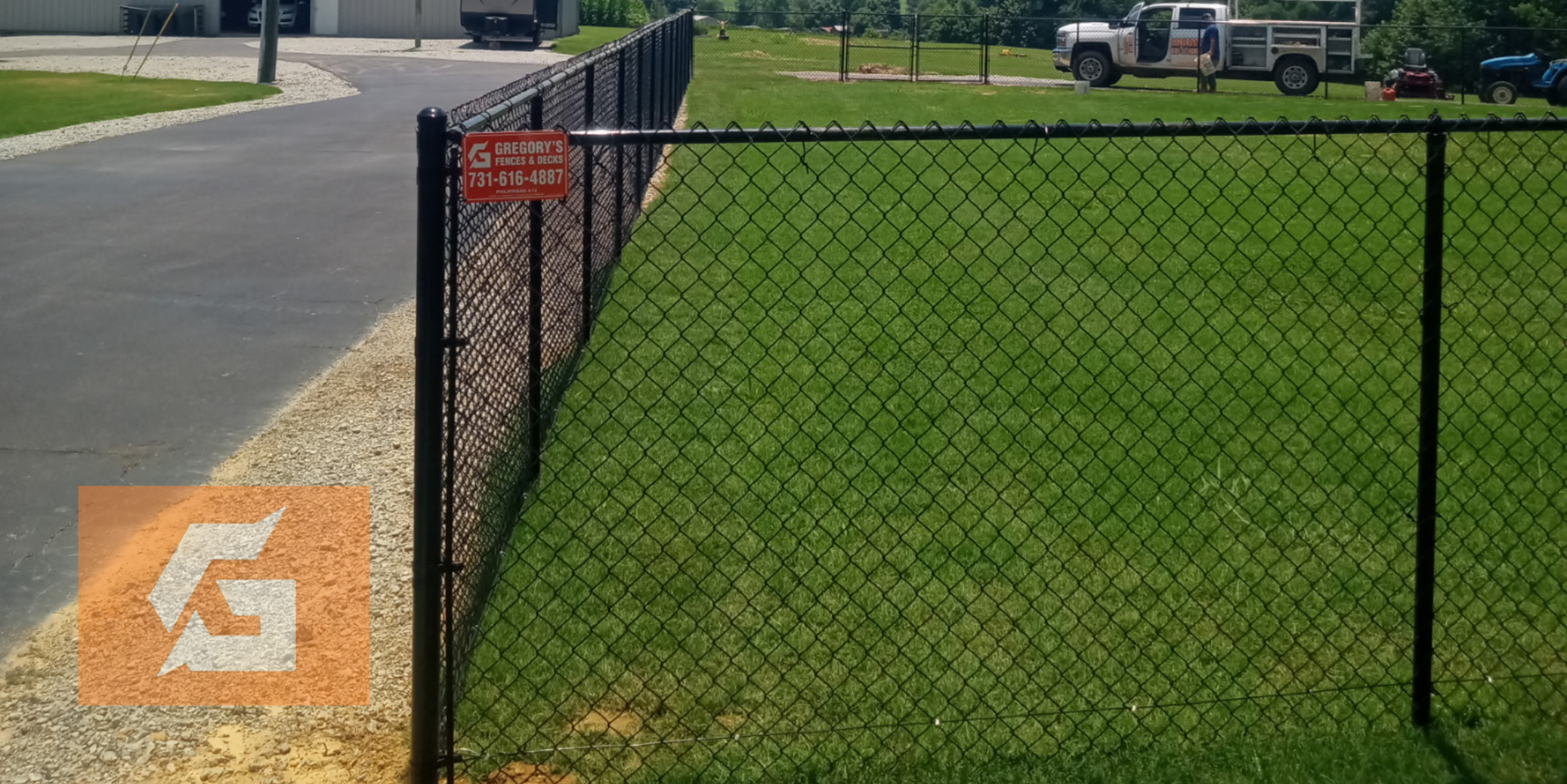 A black chain link fence borders a grassy area, with a sign and a vehicle in the distance.