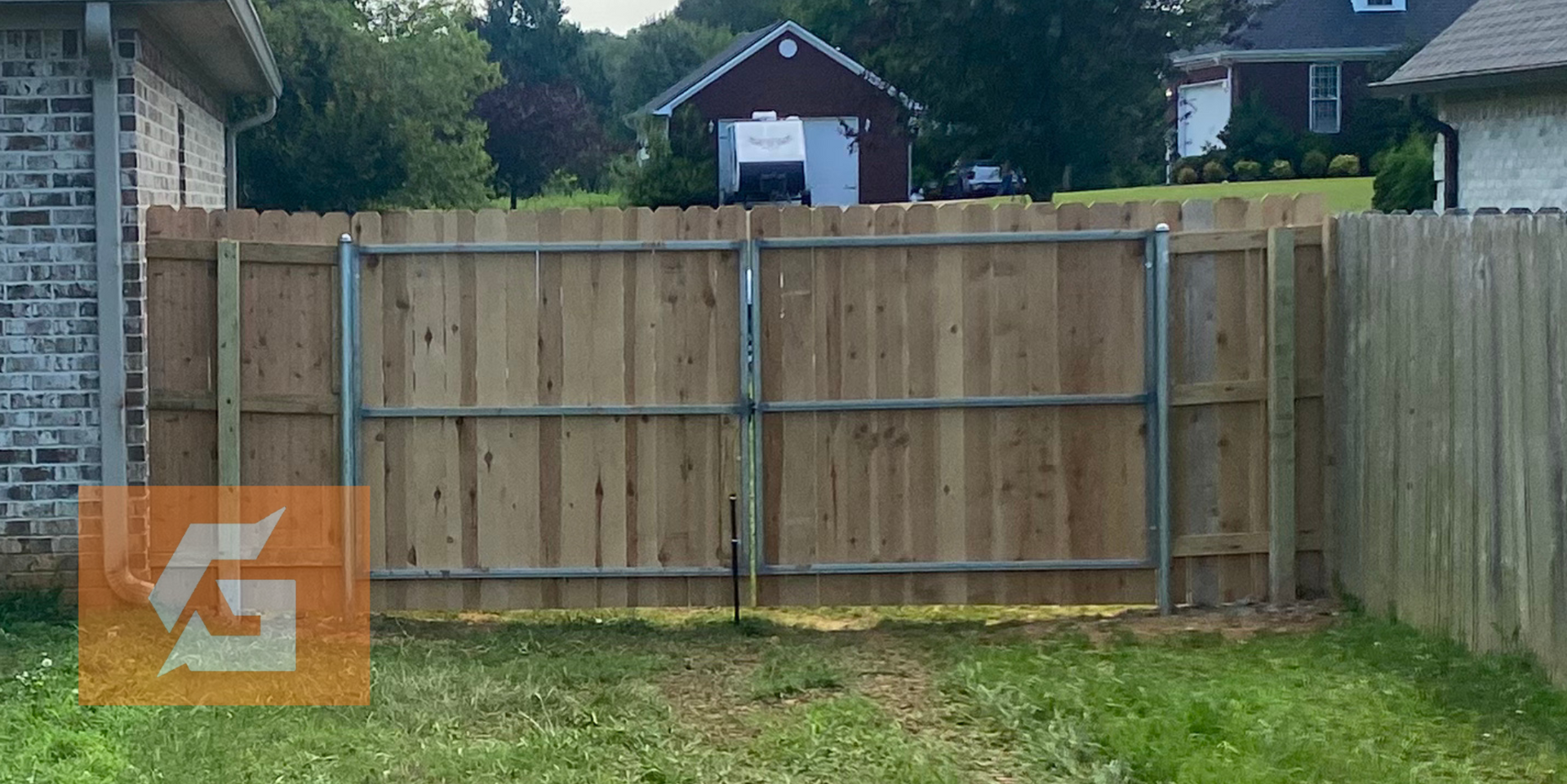 Wooden fence with a metal gate, grassy yard, two houses in the background.