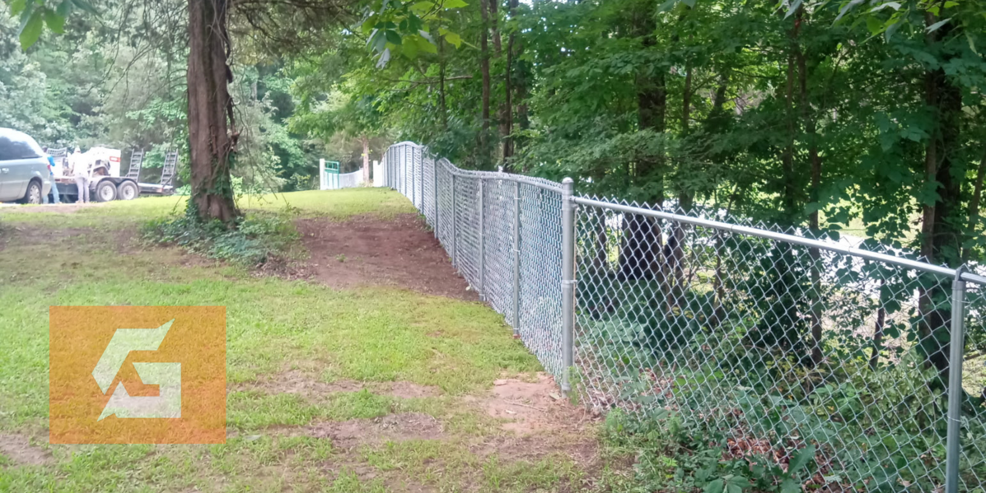 Chain-link fence beside a wooded area with a grassy yard and several vehicles visible.