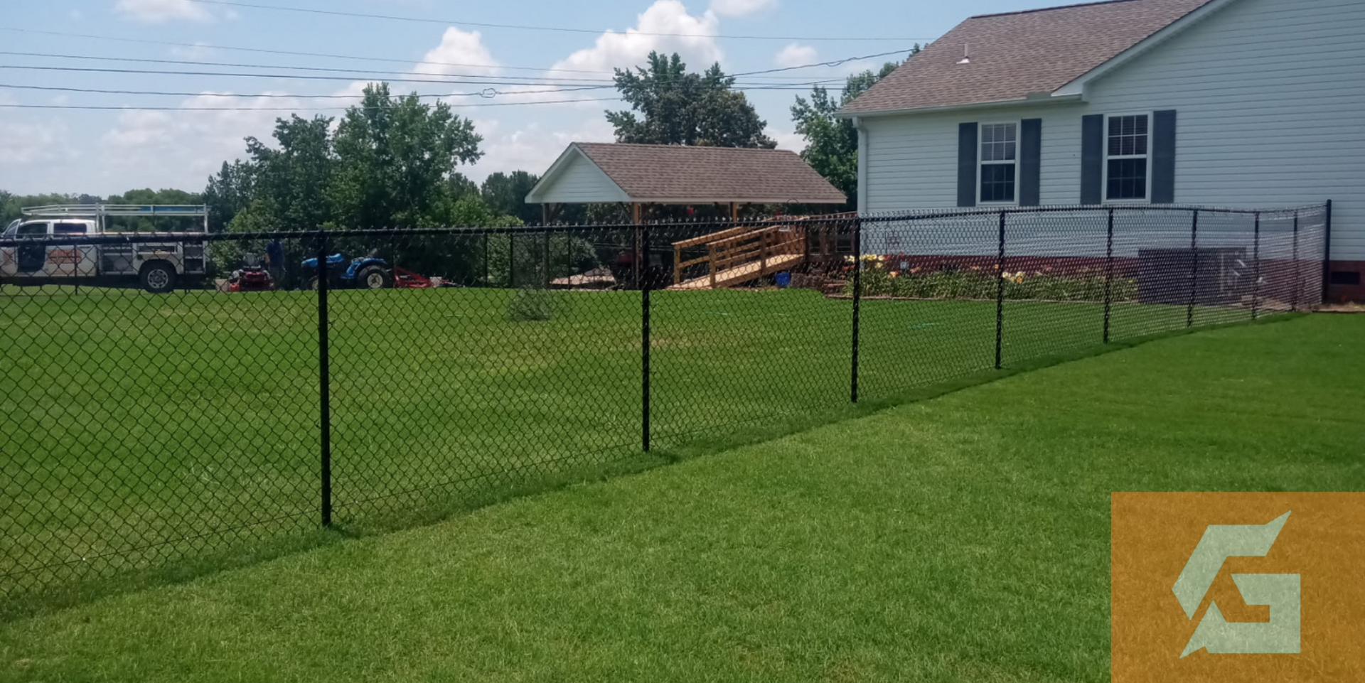 Black chain-link fence in a grassy yard, with a house and trees in the background on a sunny day.