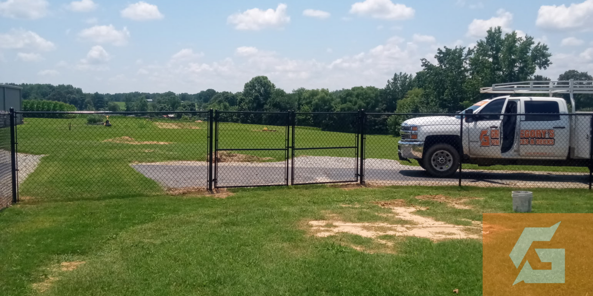 A chain-link fence encloses a grassy area with a truck parked nearby on a sunny day.