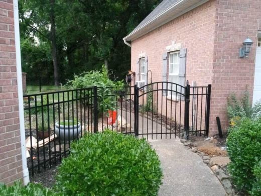 Black metal fence and gate leading to a brick house, with green shrubs and a walkway.