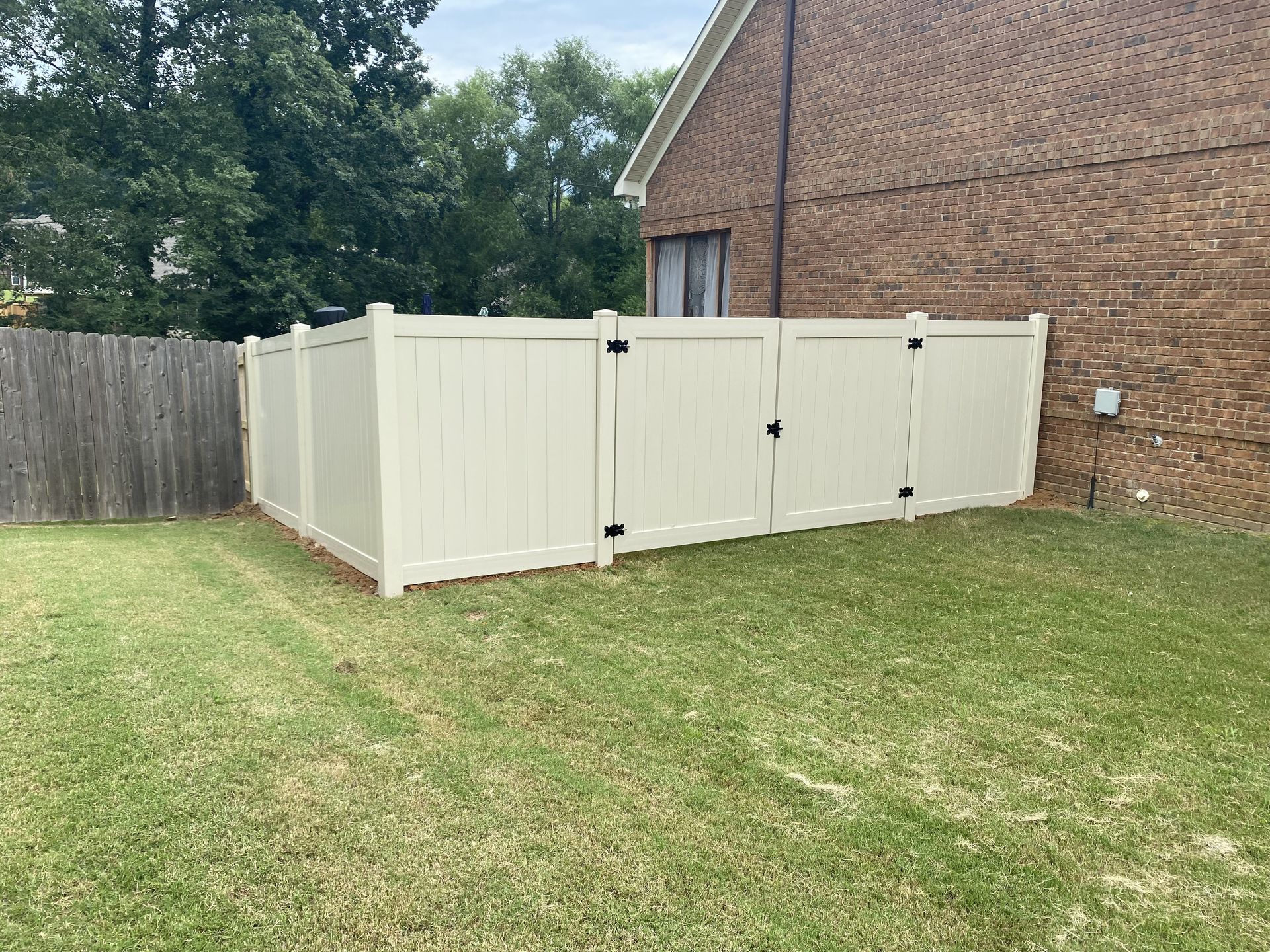 Cream-colored vinyl fence with gate next to a brick building. Green grass in front.