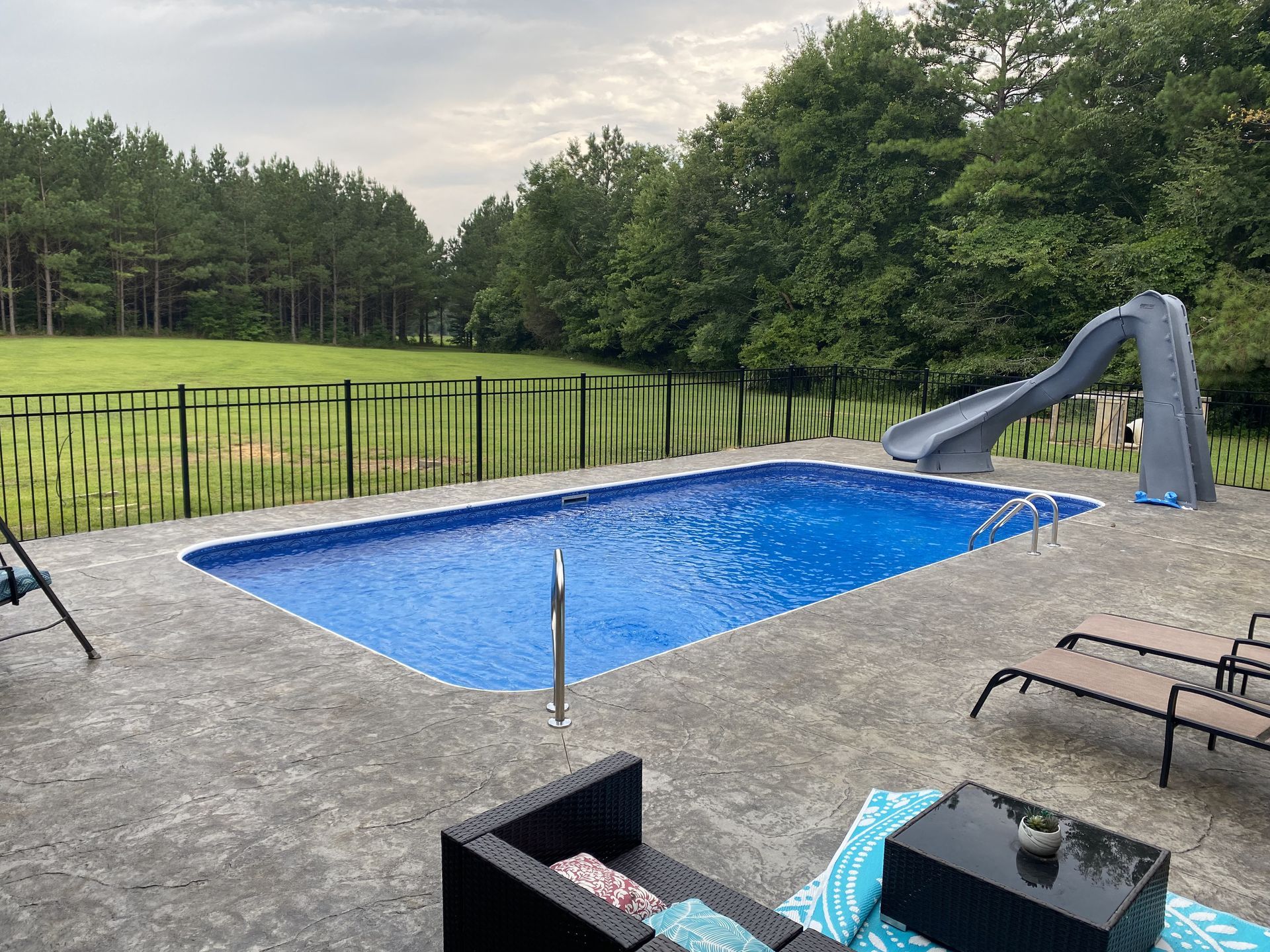 Rectangular pool with a slide, surrounded by a concrete patio, lawn, trees, and a black fence.