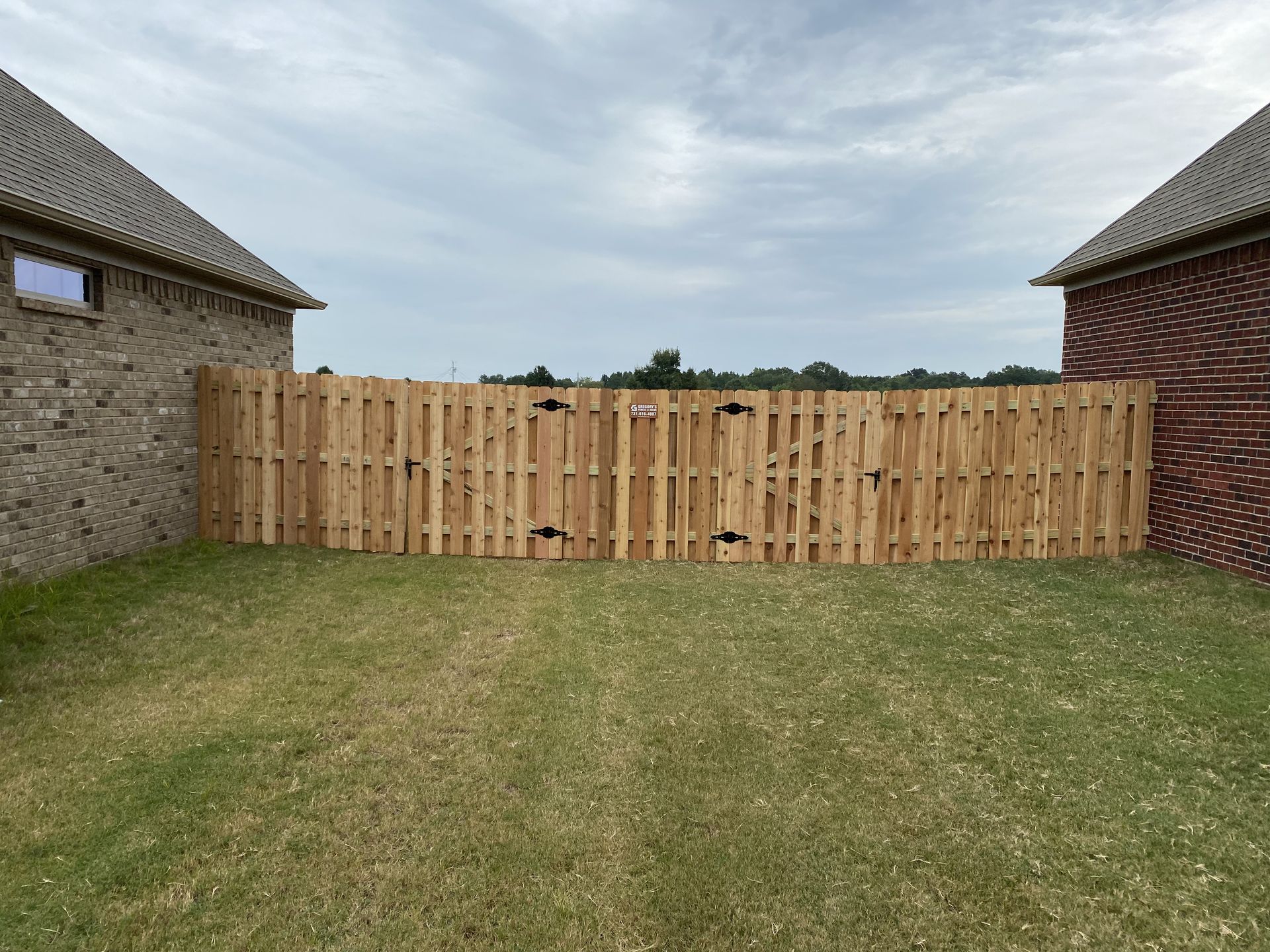 A wooden fence separates two brick houses in a grassy backyard under a cloudy sky.