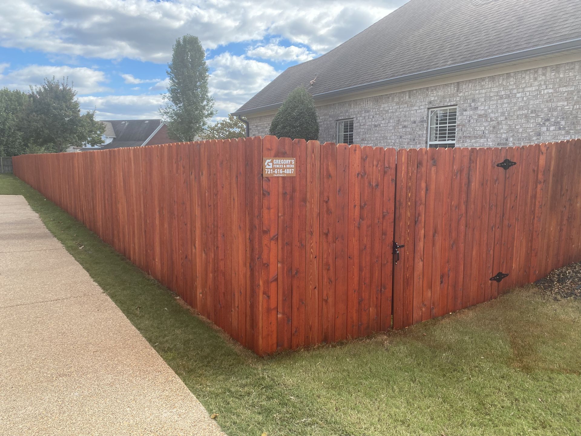 Red-stained wooden fence surrounds a house, with a gate, sidewalk, and green lawn.