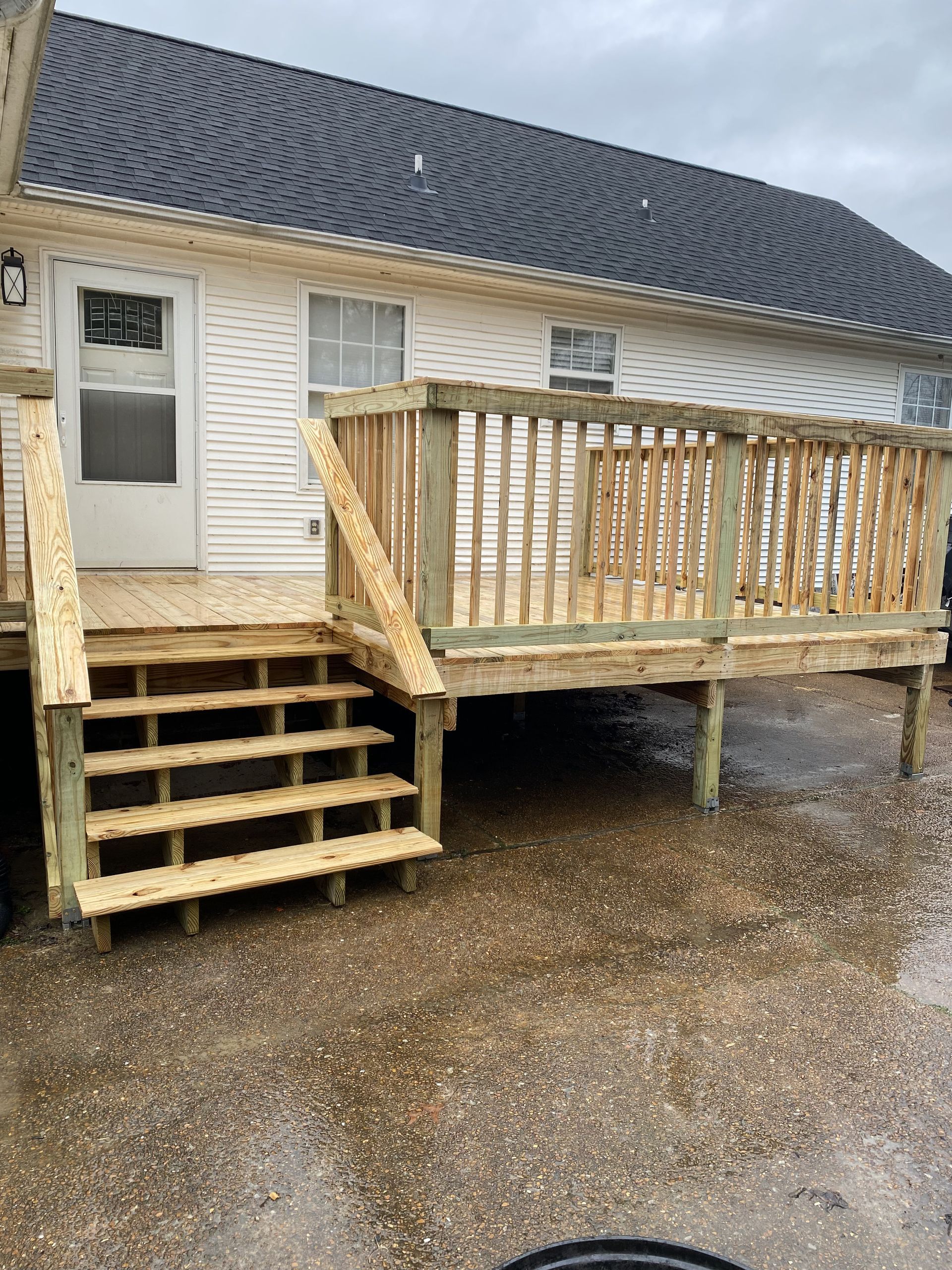 New wooden deck with stairs, attached to a white house with a black roof. Overcast day.
