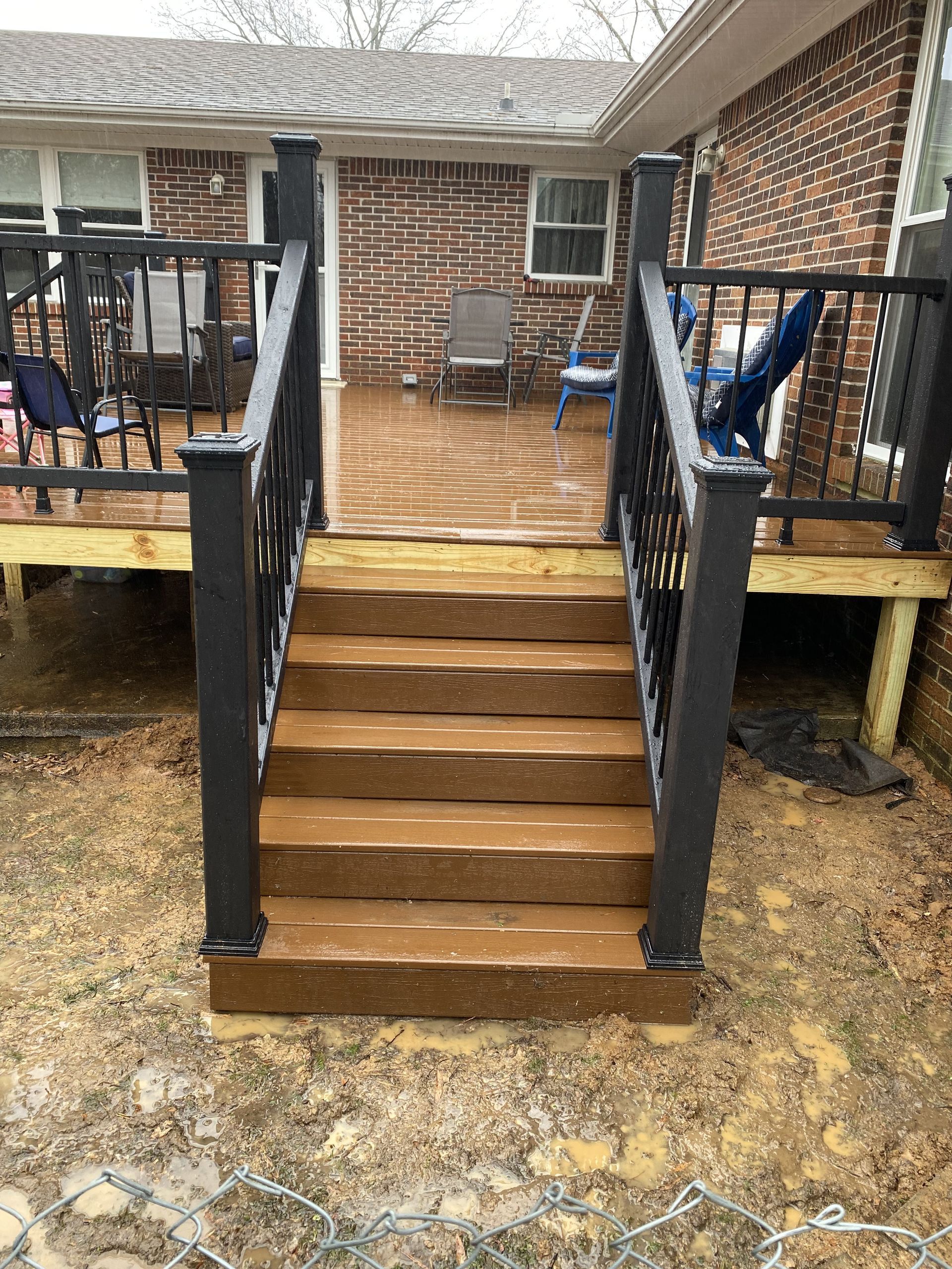 Brown deck with steps, black railing, and brick house background.