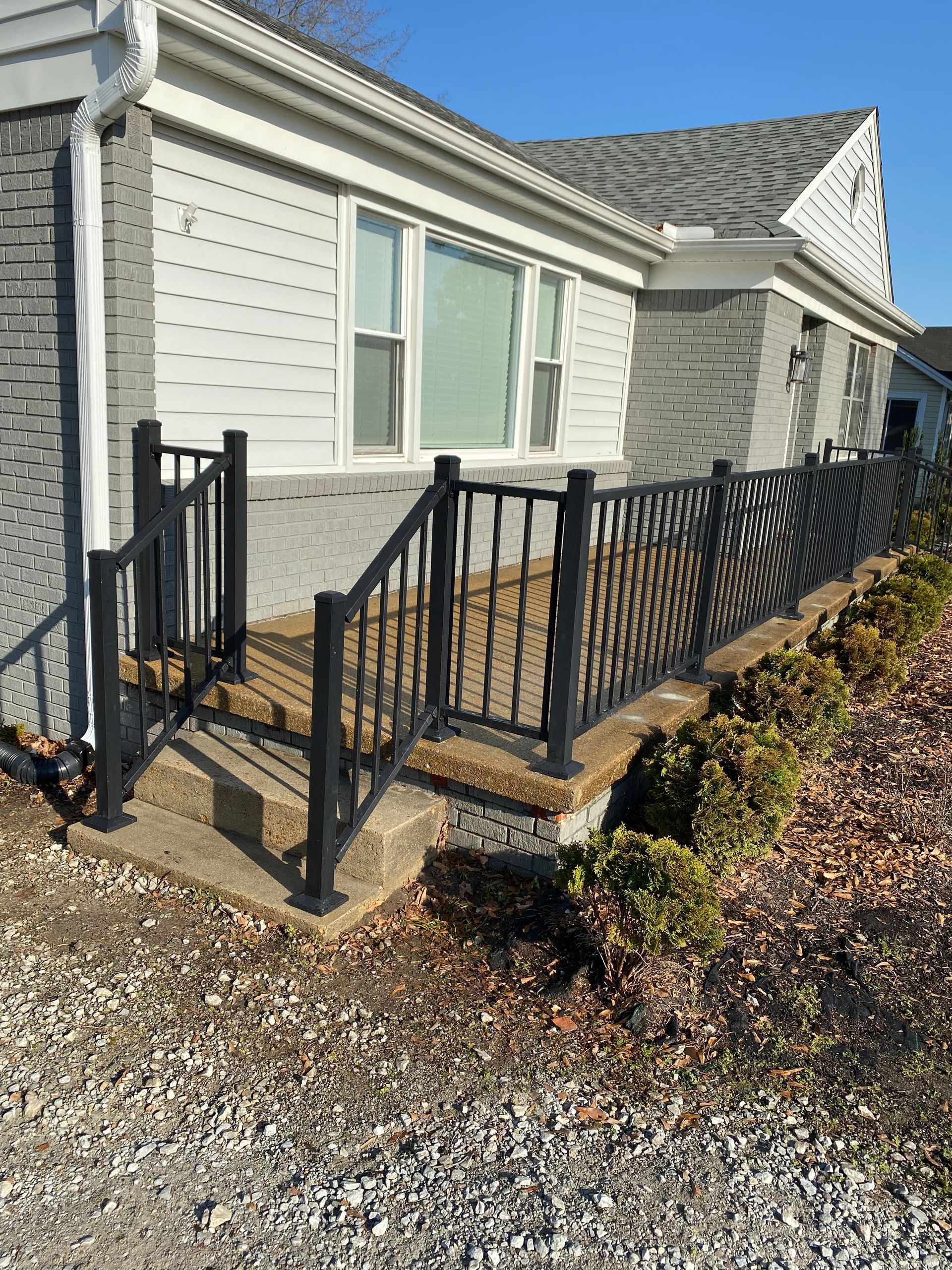 Black railing surrounds a porch with a light-colored house, small bushes and gravel.
