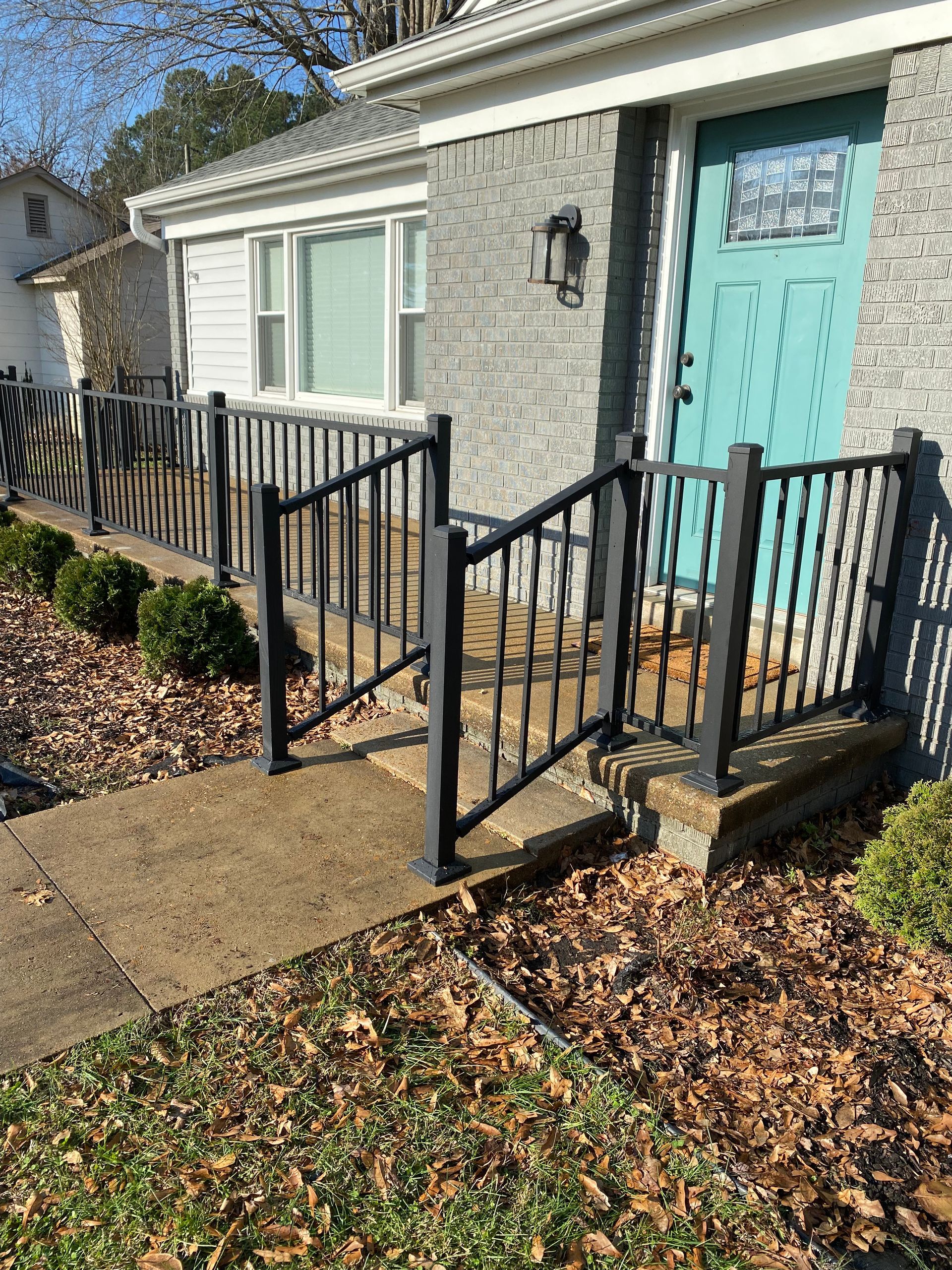 Gray house with teal door and black railing over a concrete walkway with fall leaves.