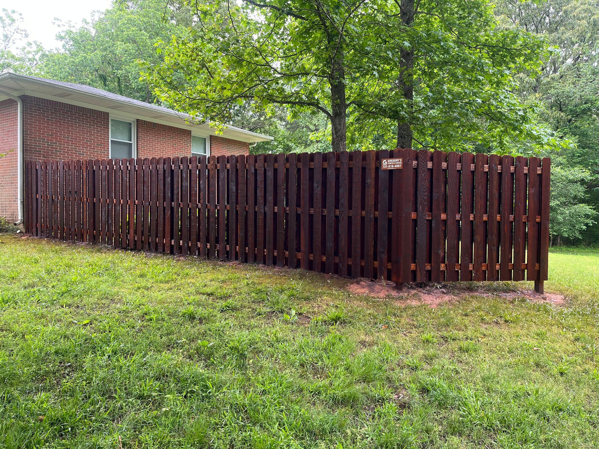 Brown wooden fence along a brick house and green grassy yard.