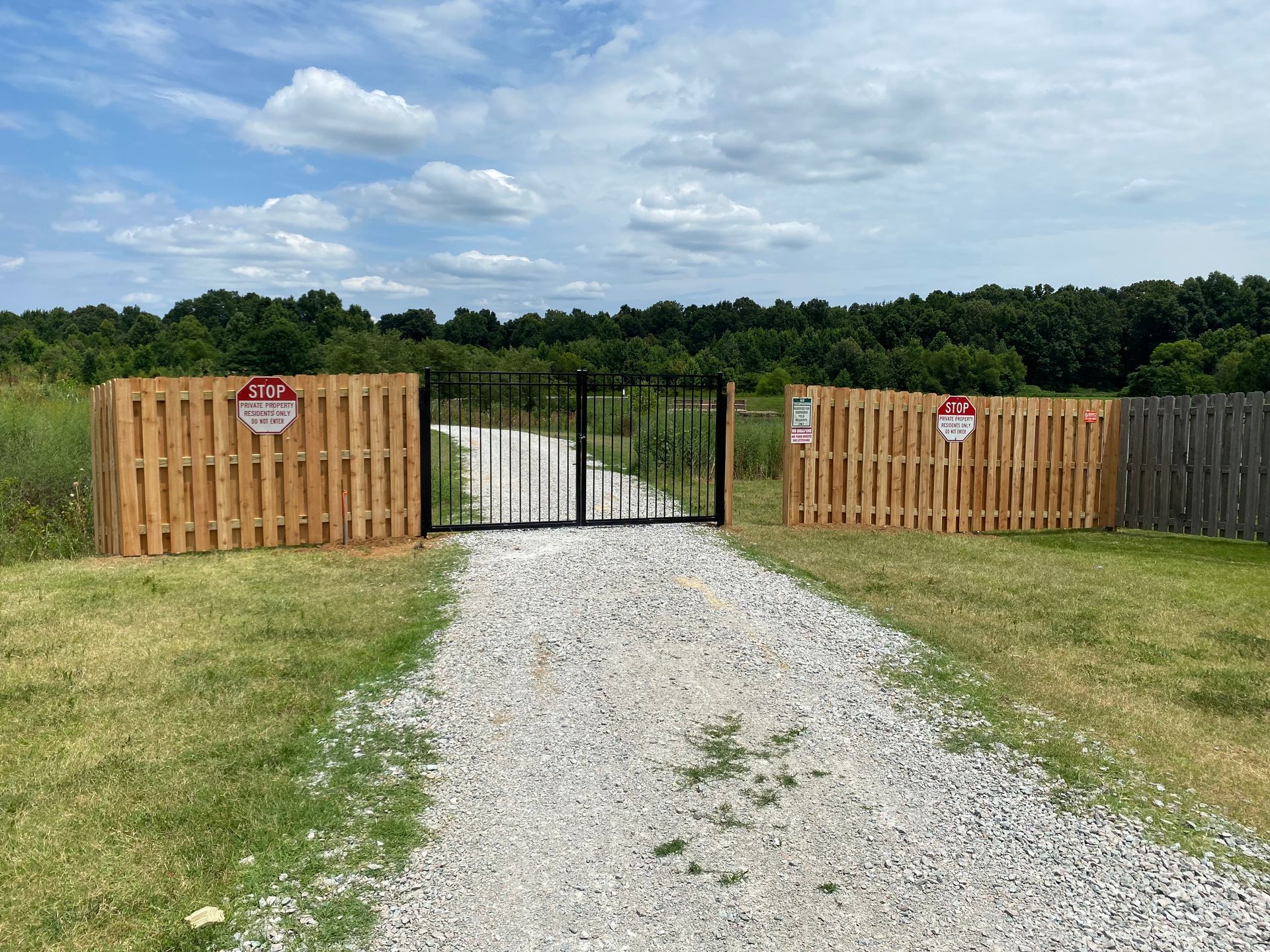 Gravel path leads to a black gate, flanked by wooden fences with signs, in a field with trees under a cloudy sky.