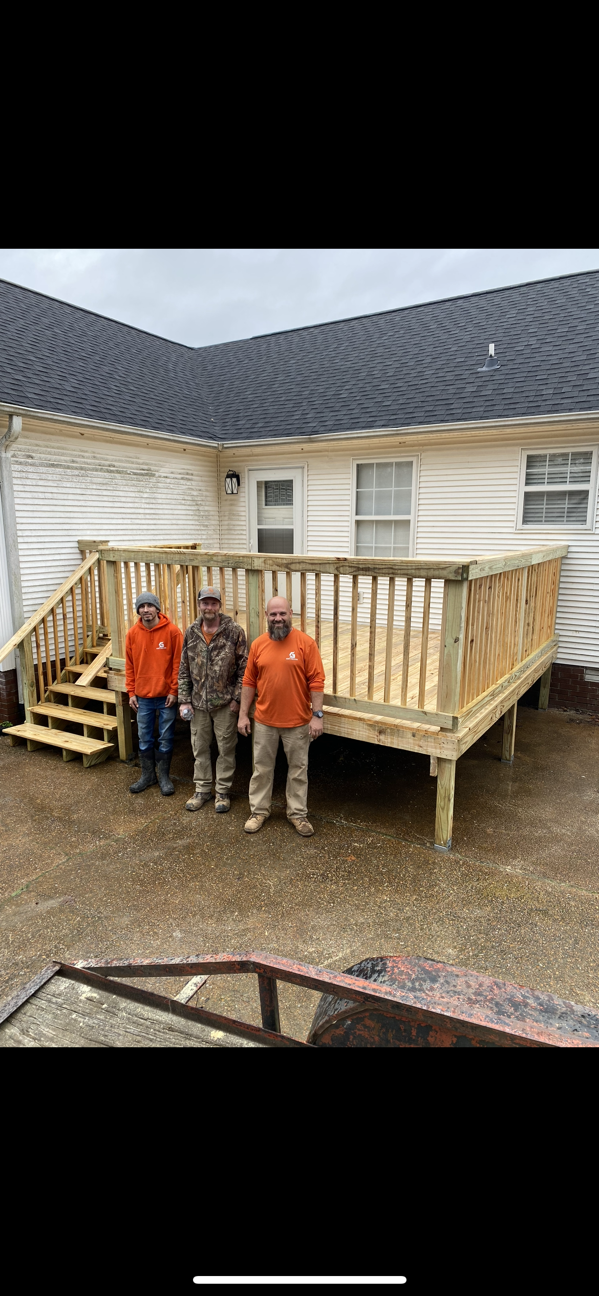 Three men stand on a newly constructed wooden deck in front of a house. Cloudy day.