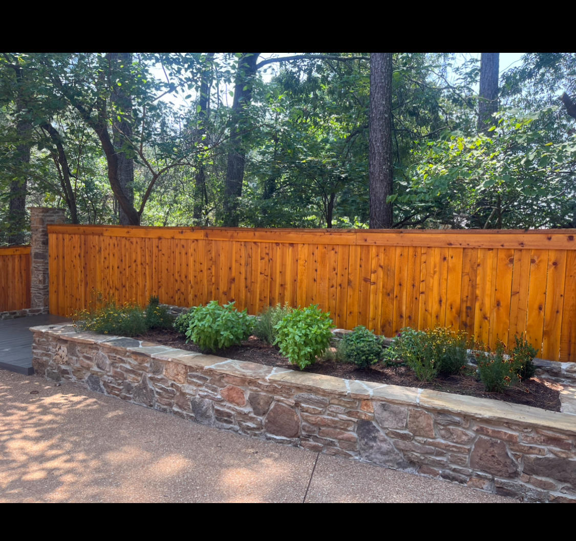 A cedar wood fence sits behind a stone-walled garden with bushes, plants, and a forest backdrop.