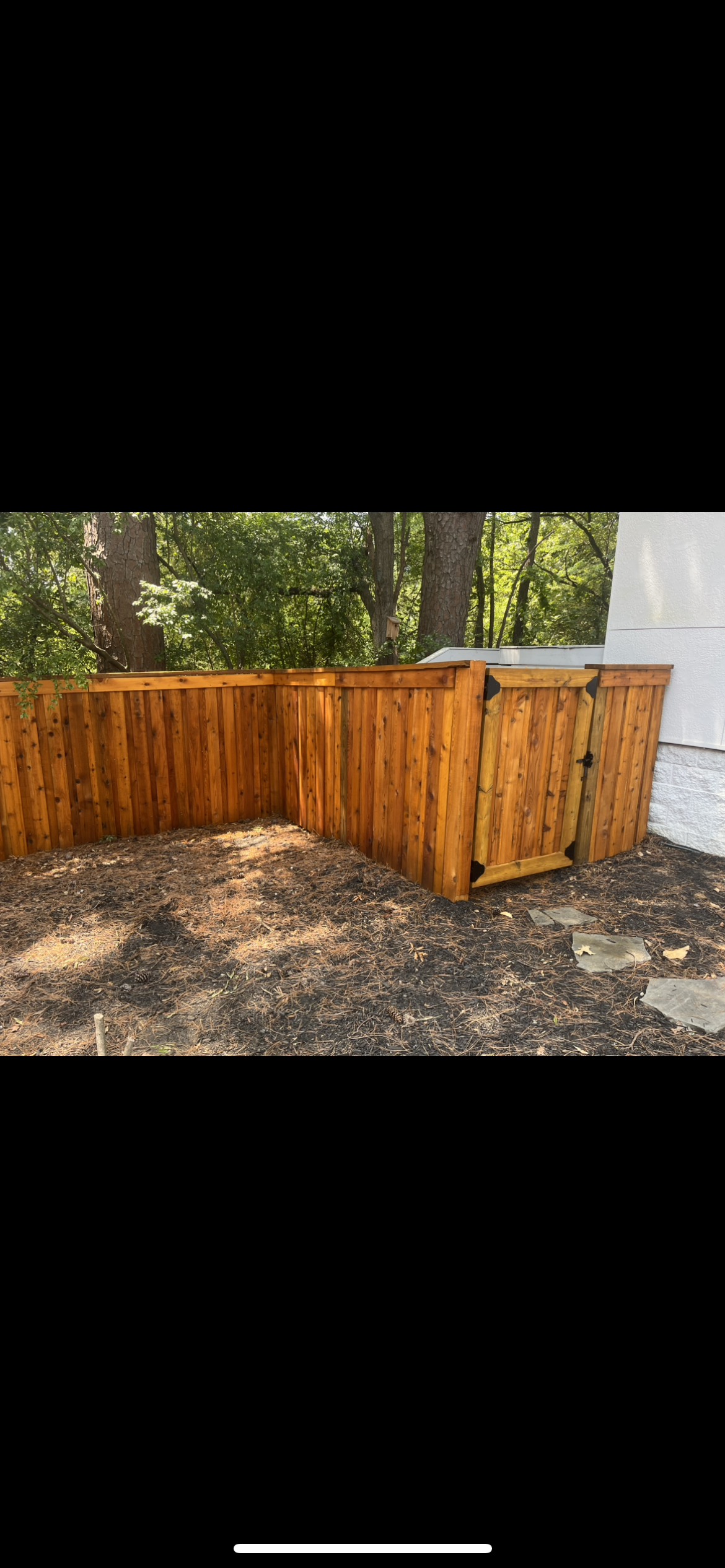 Wooden fence and storage bins in front of a wooded area. Brown wood, gravel, and green trees.