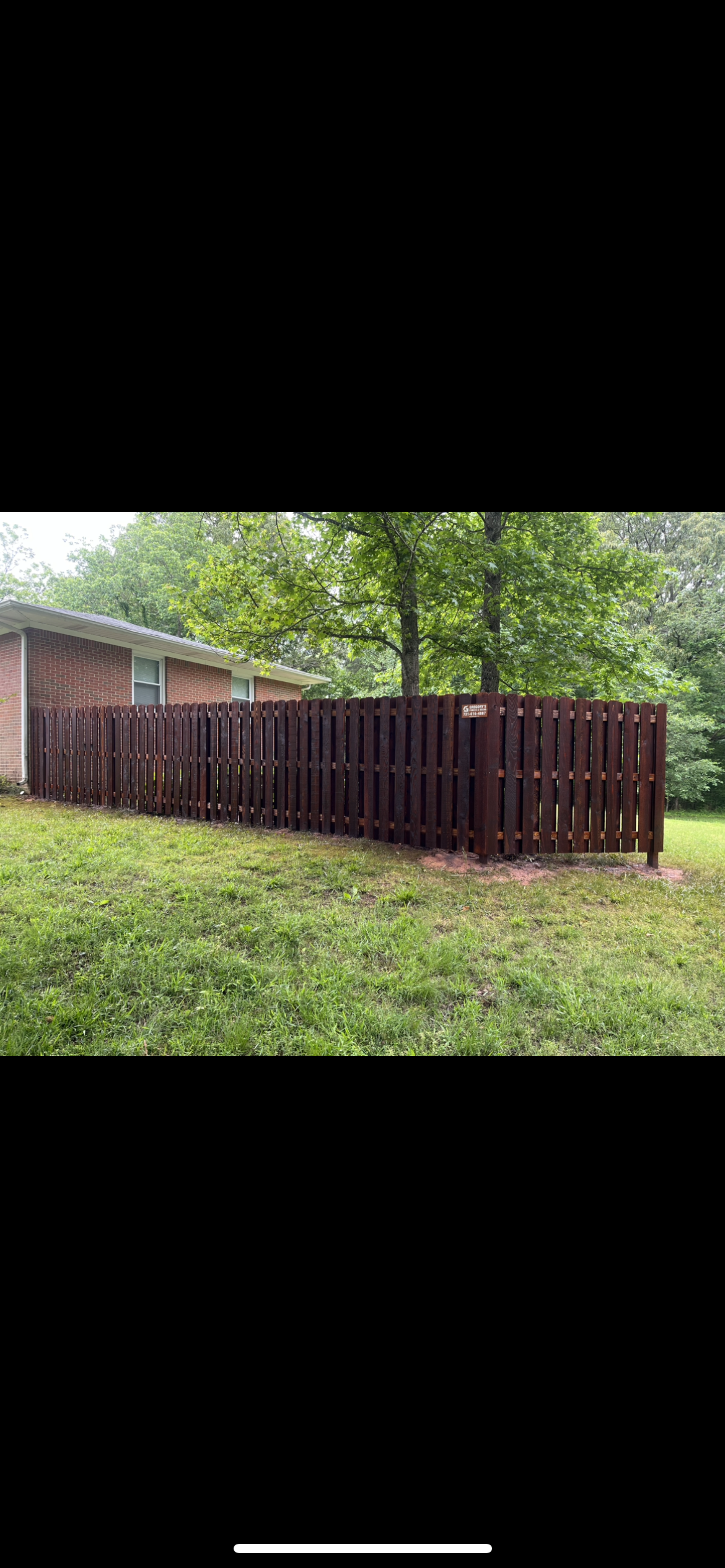 A brown wooden fence surrounds a grassy yard, trees and a house in the background.
