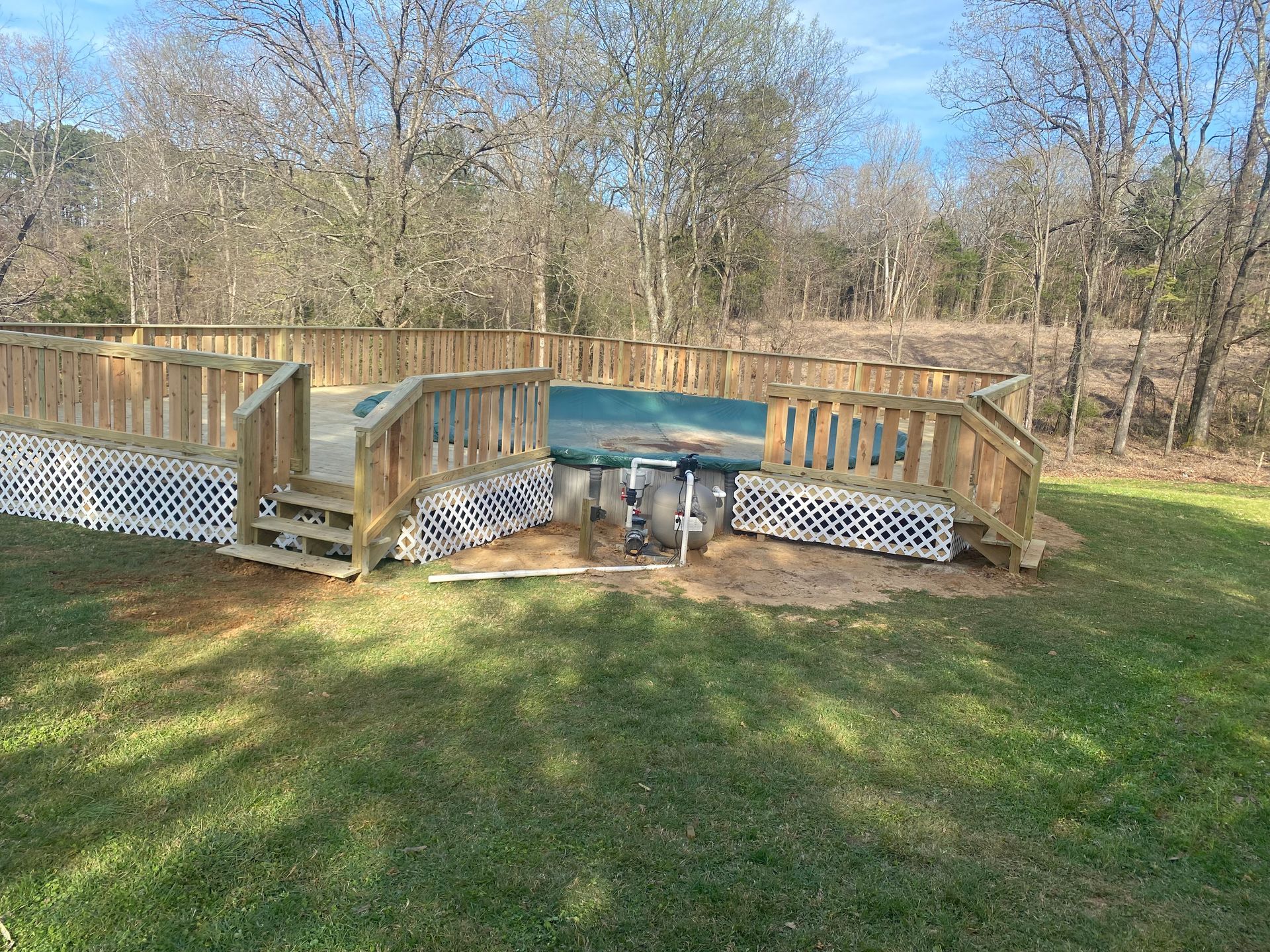 Wooden deck with stairs, against a backdrop of trees. White latticework and a water feature.