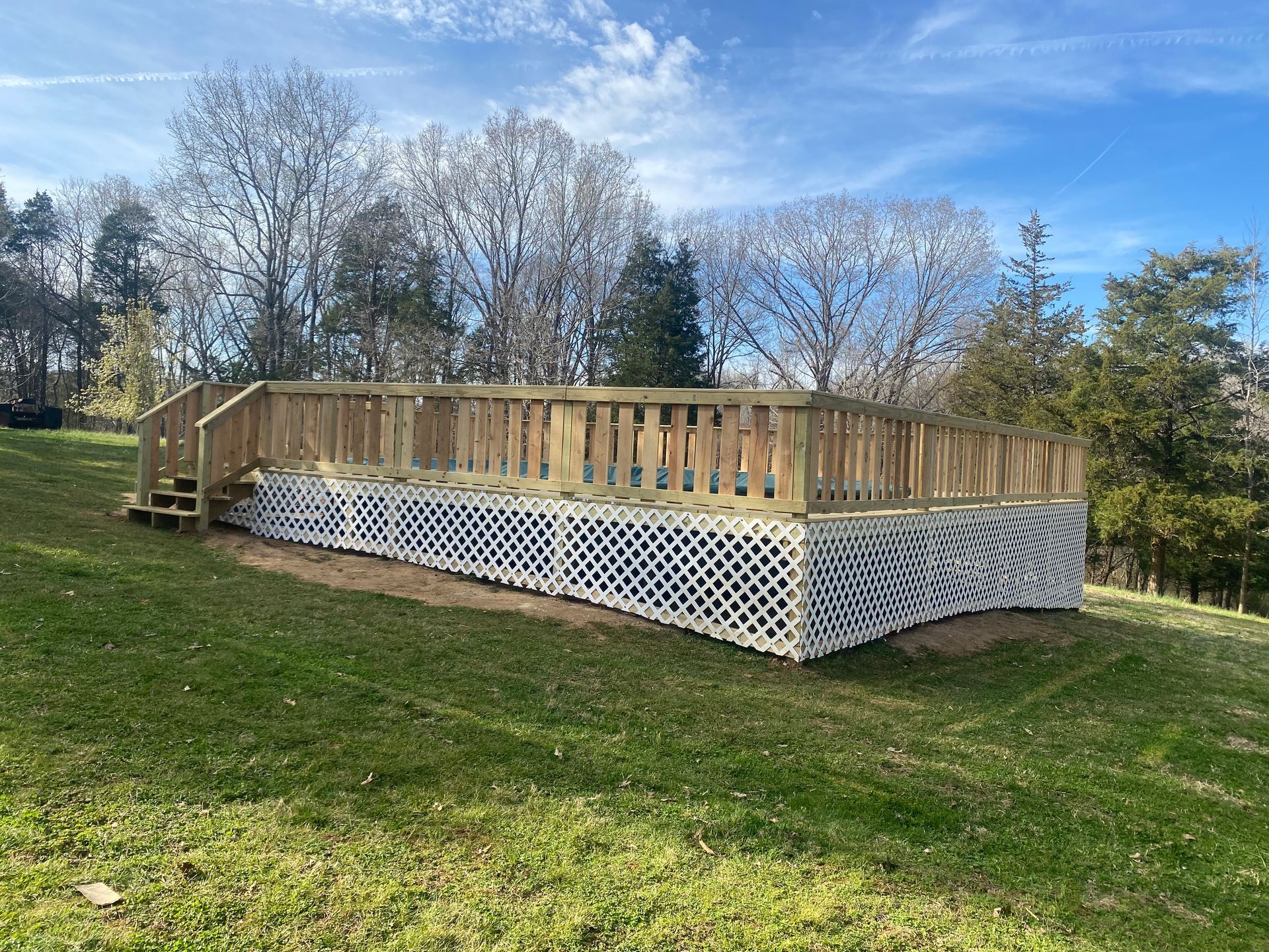Wooden deck with lattice skirting on a grassy hill, surrounded by trees under a blue sky.