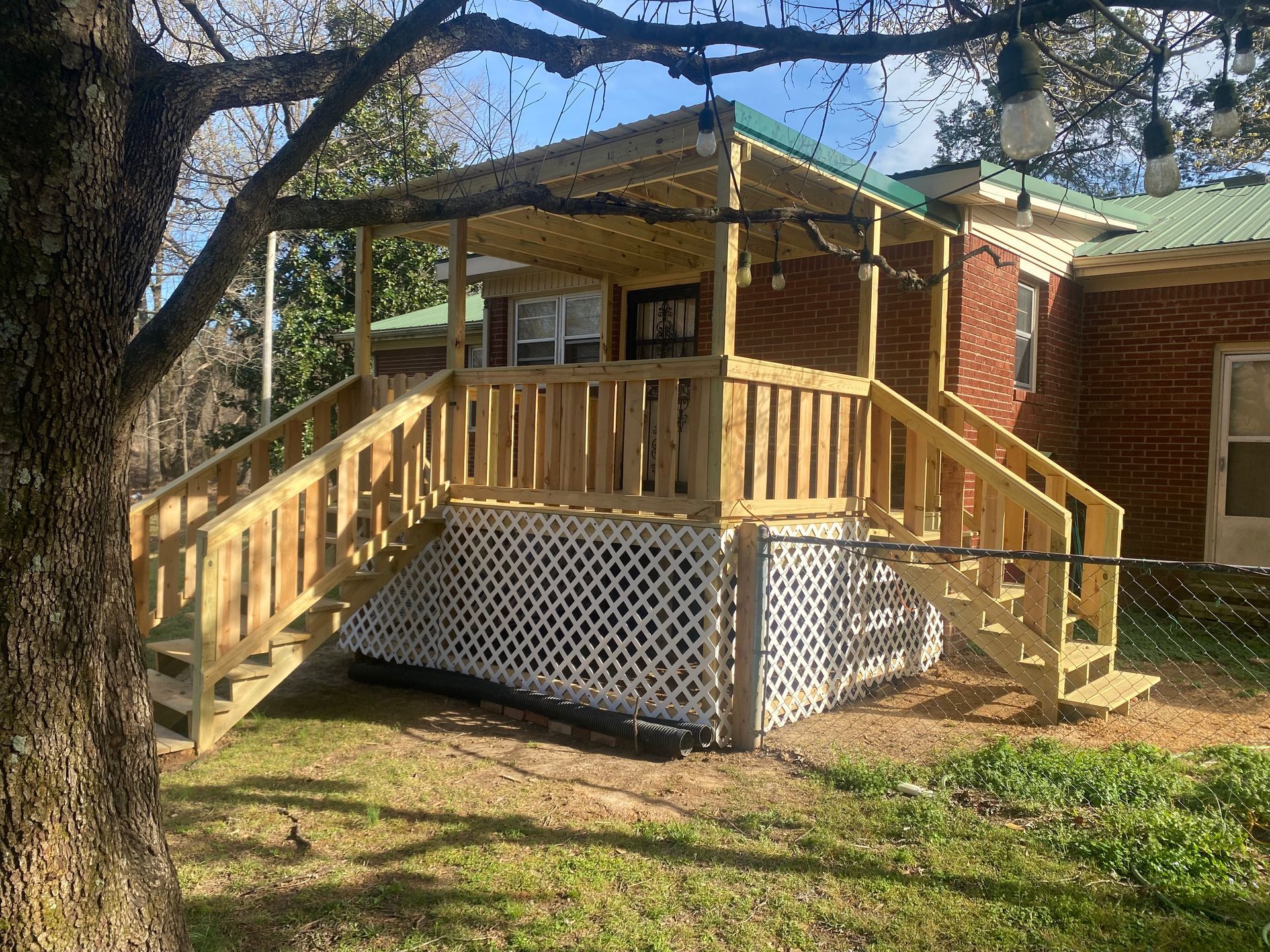 Wooden deck with stairs attached to a brick house, lattice siding, and hanging lights under a tree.