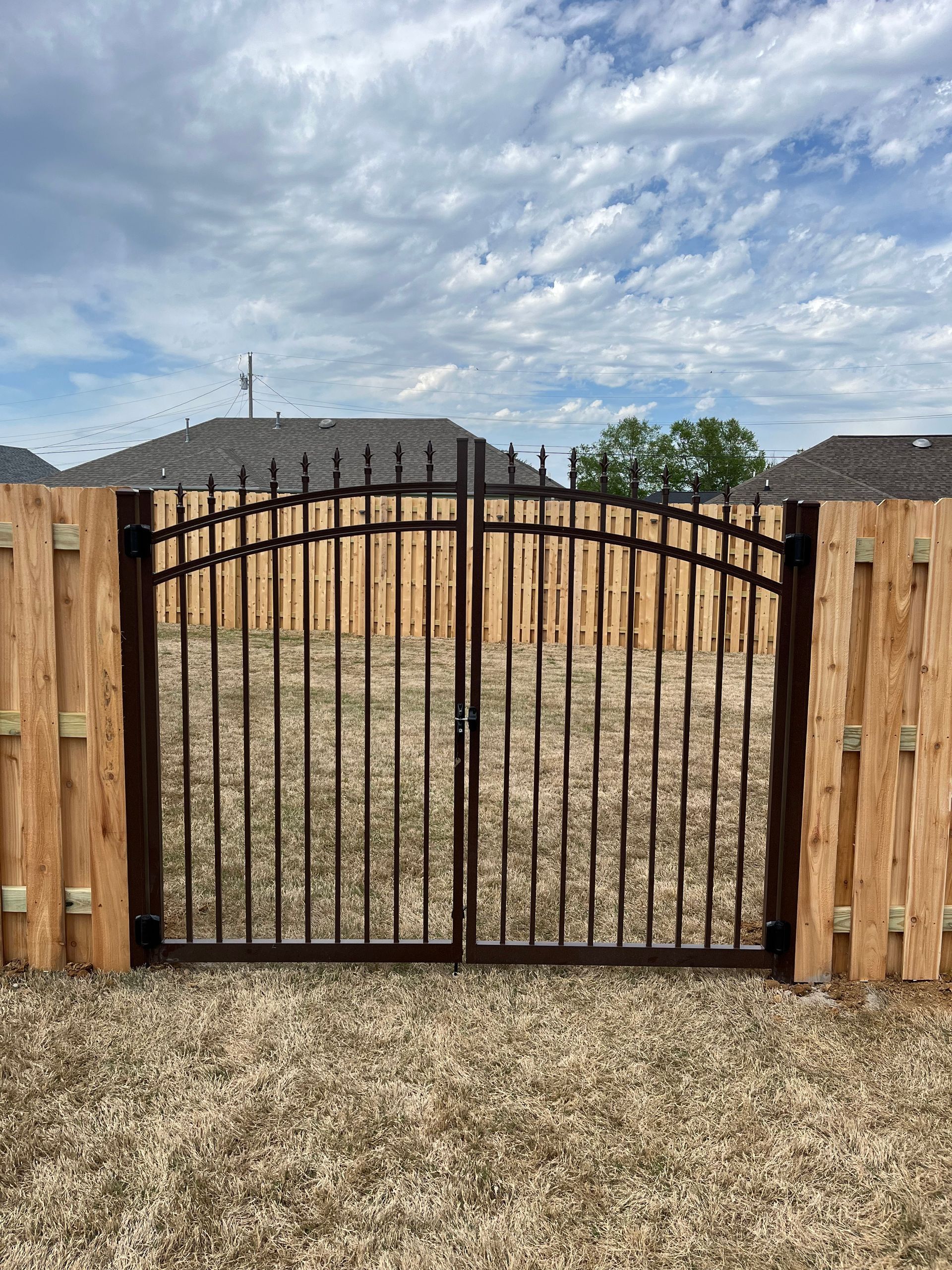 Black wrought-iron gate between wooden fence posts against a cloudy sky.