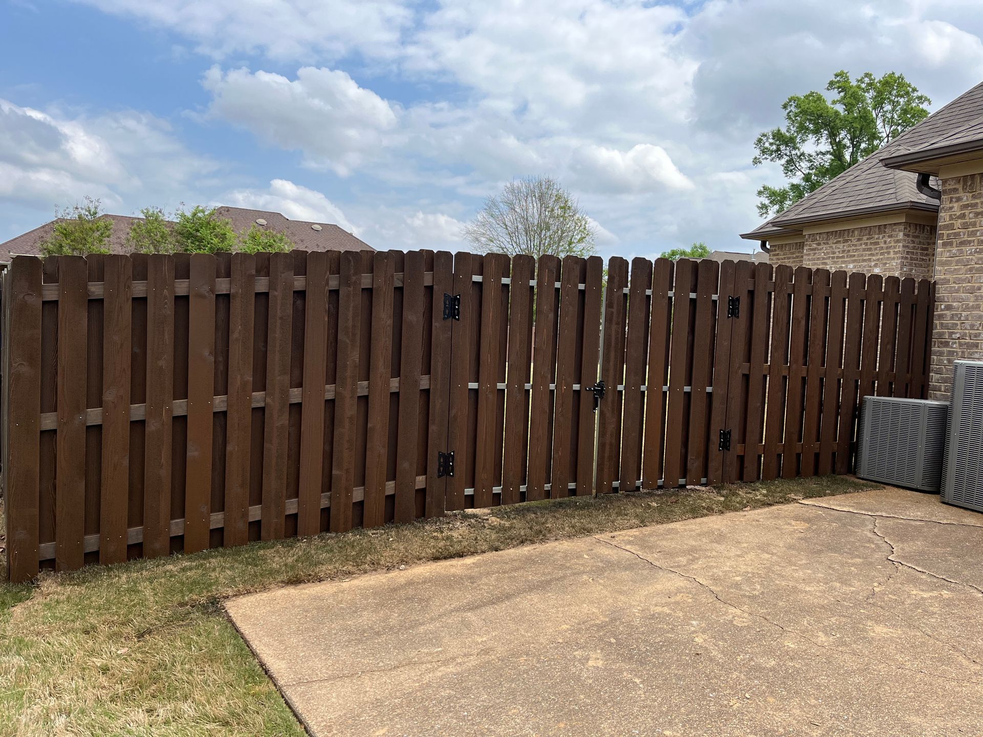 Brown wooden privacy fence in a backyard, against a brick house under a cloudy sky.