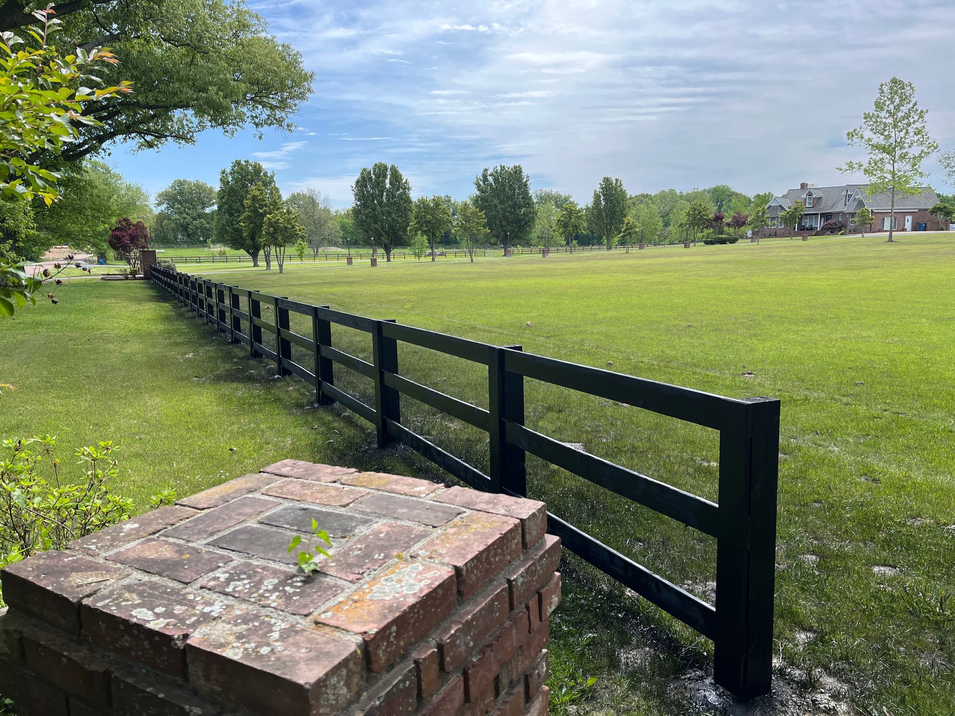 Black fence bordering a green field under a cloudy sky, with a brick post in the foreground.