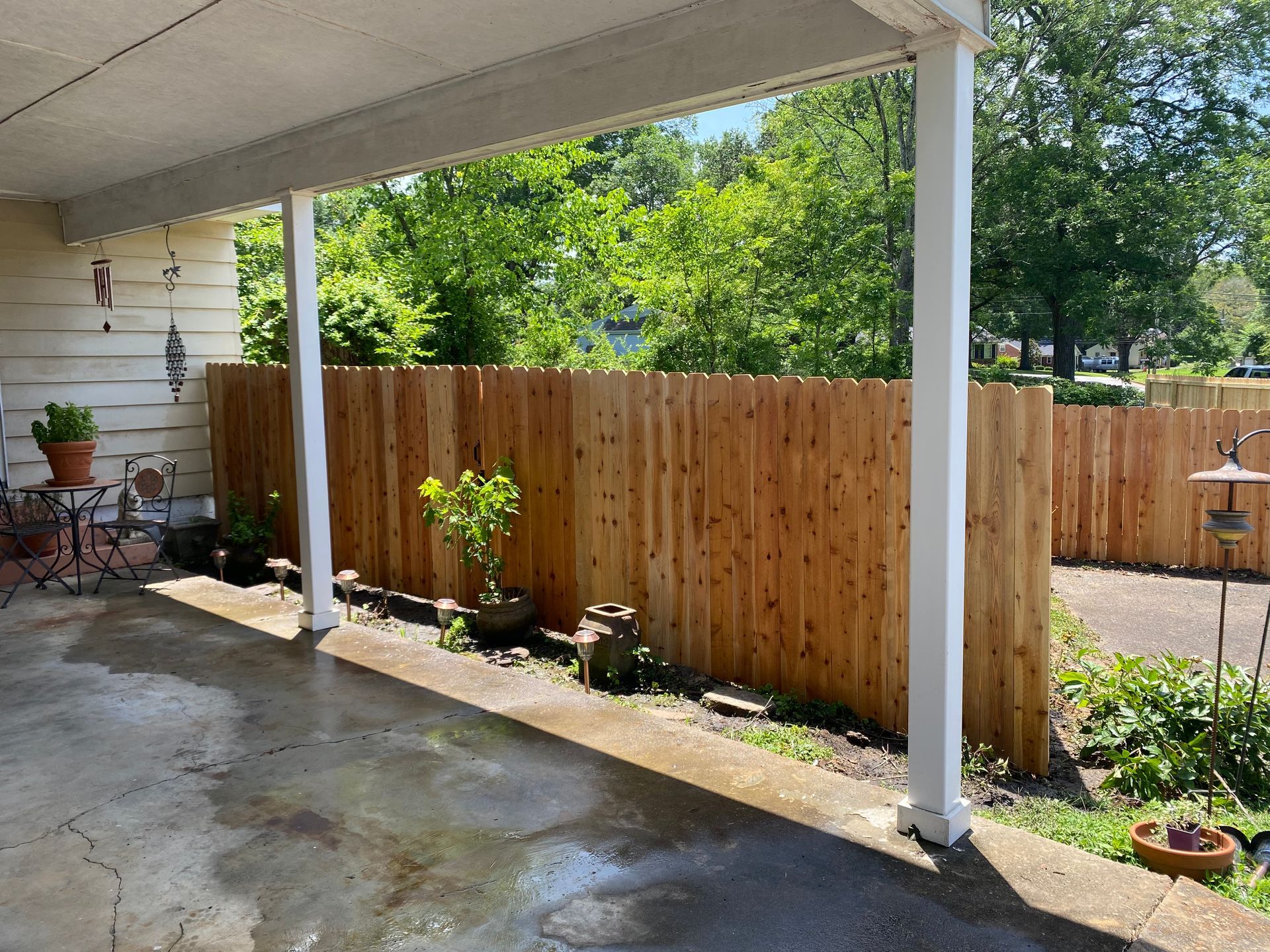 Covered porch with wet concrete floor, wooden fence, potted plants, and surrounding trees.