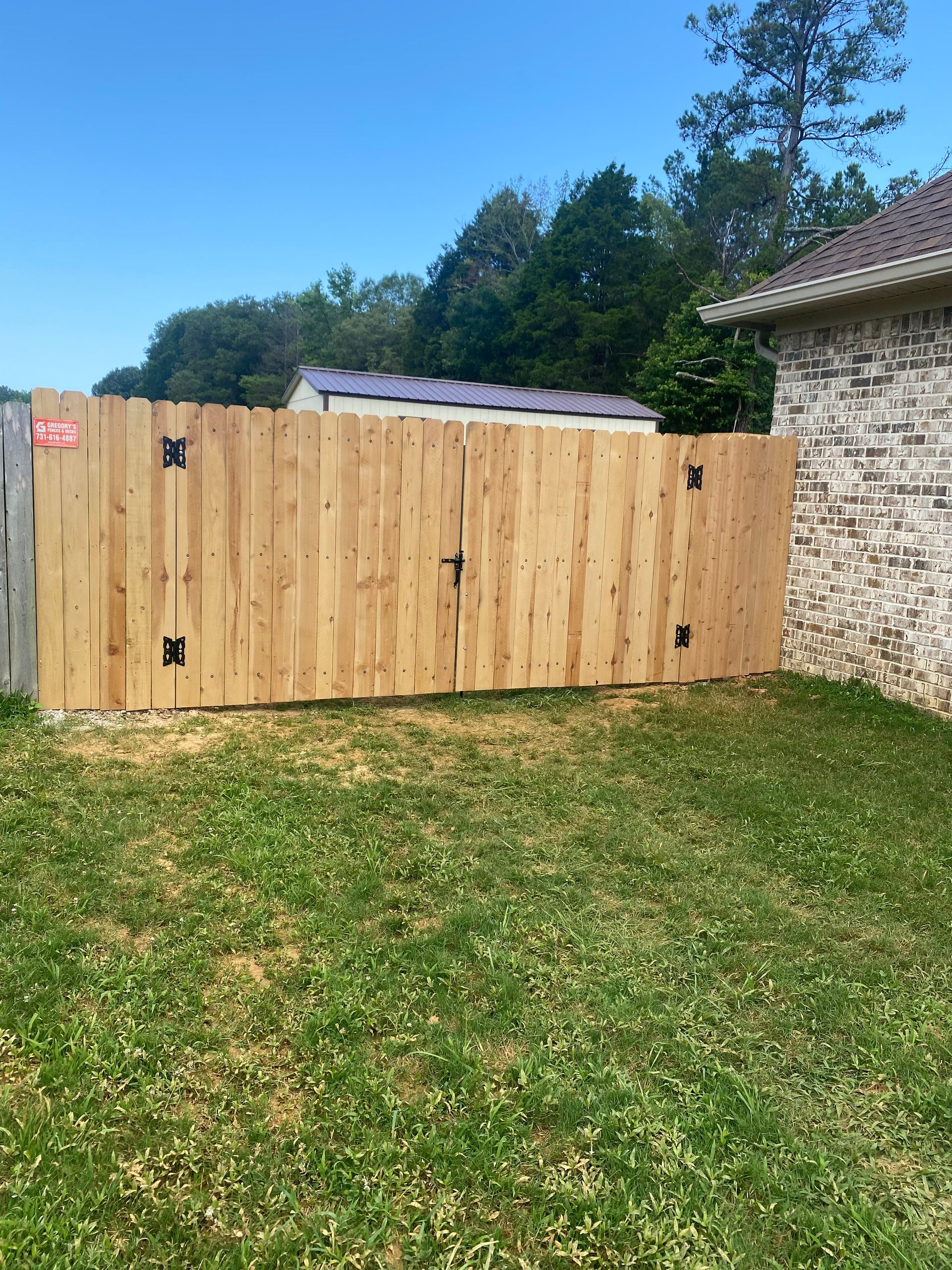 Wooden gate with black hinges and latch in a grassy yard next to a brick wall.