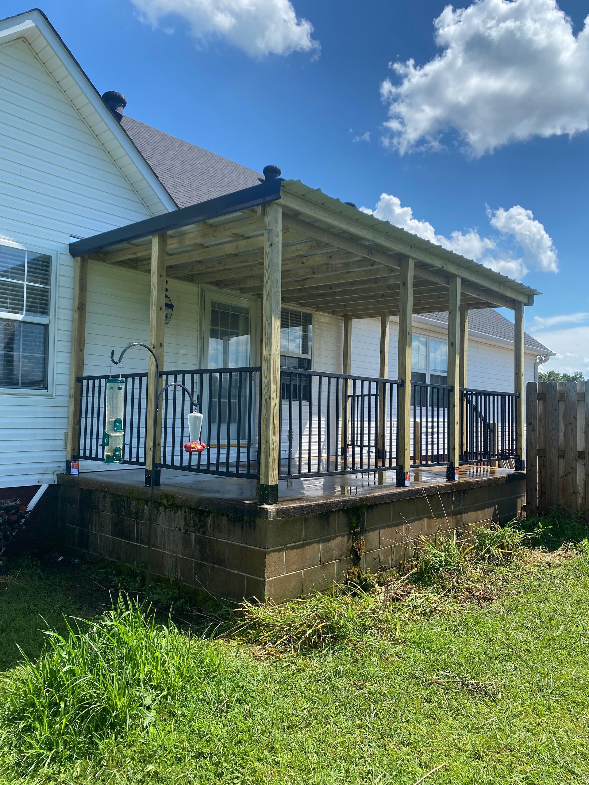 Covered porch with black railings and wooden supports attached to a white house, green grass in front.
