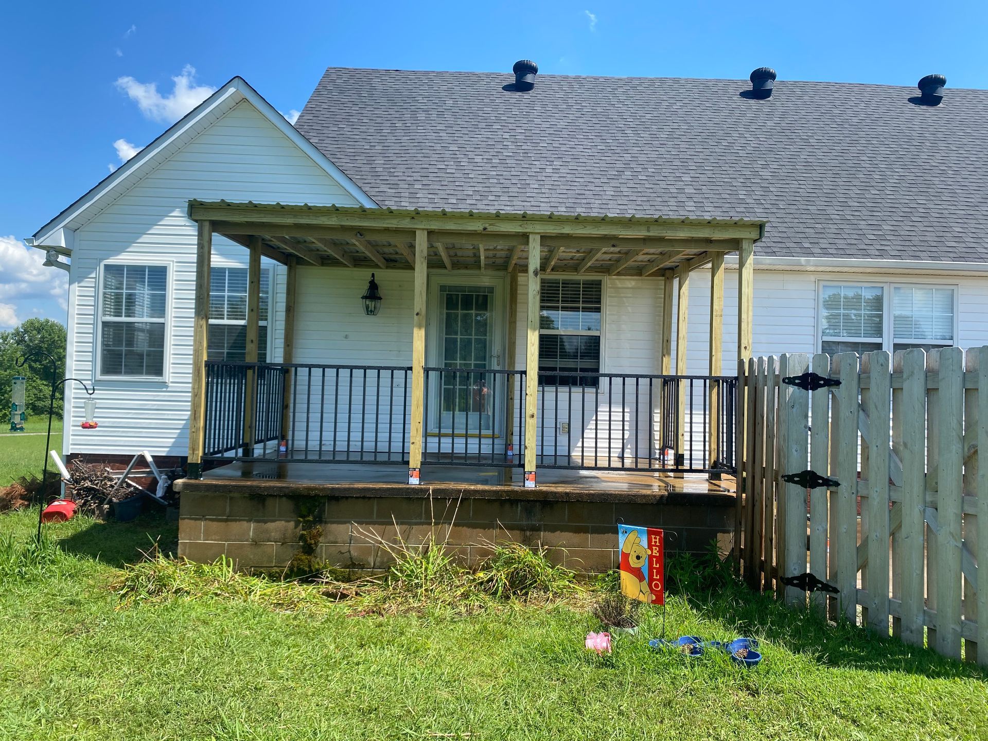 White house with a small porch. Black railing, wooden posts, and gray roof. Green grass and blue sky.
