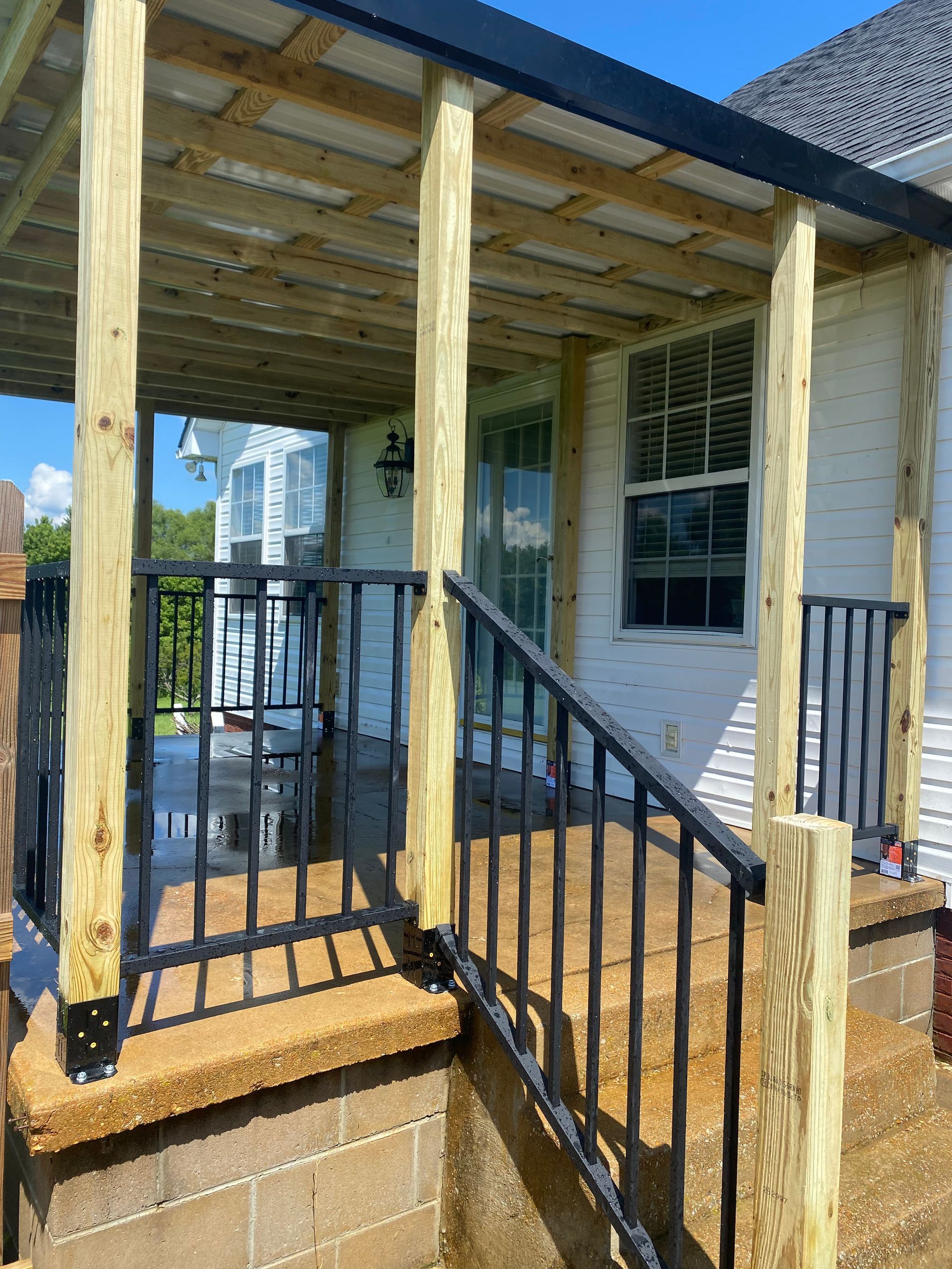 A porch with a black railing, concrete steps, and wooden posts; a white house in the background.