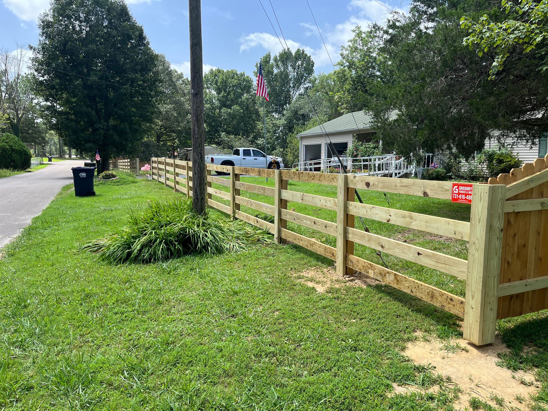 Wooden fence along a green grassy lawn near a road. White pickup truck and house in the background.