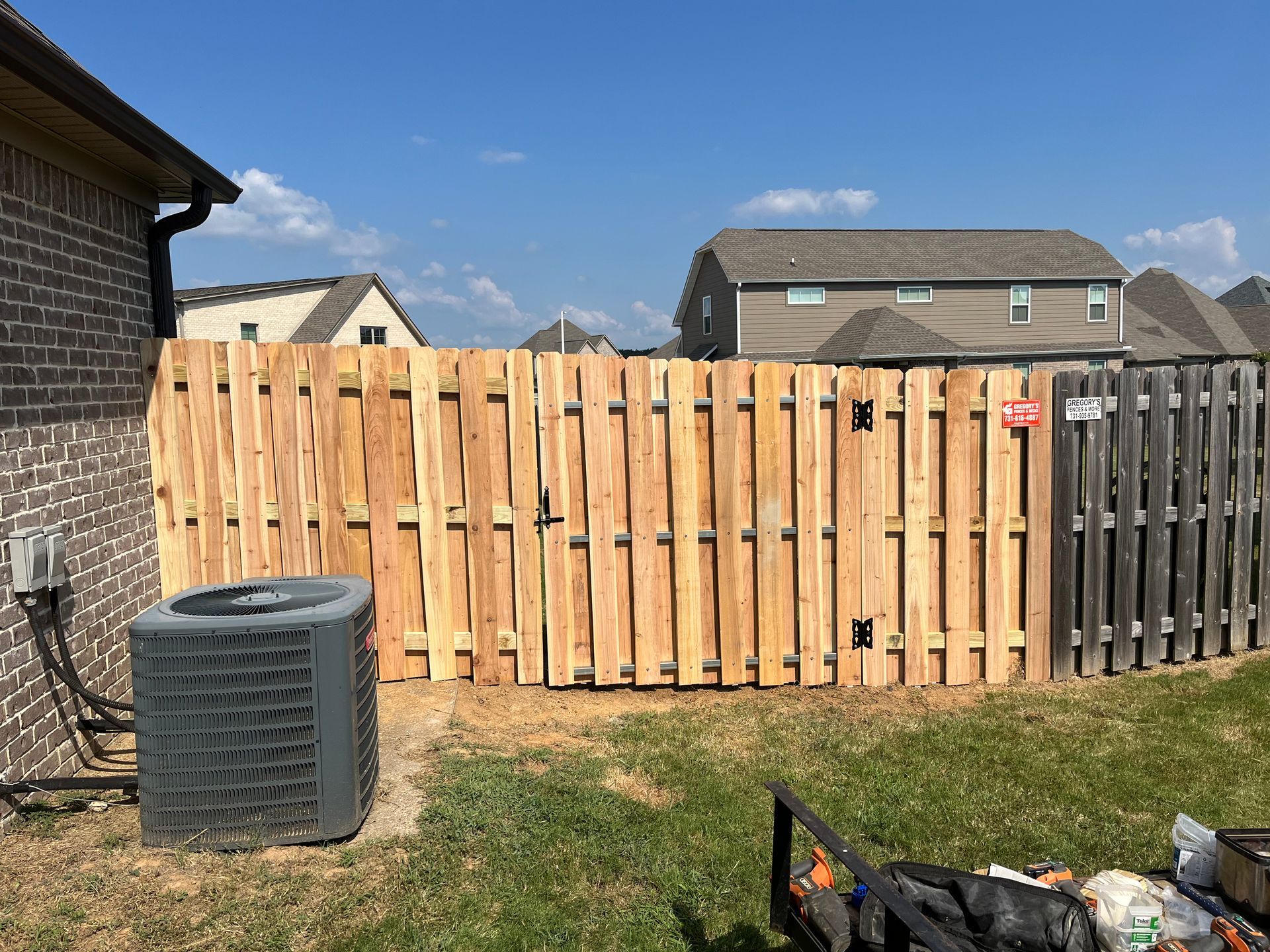 A new light brown wooden fence alongside a darker gray fence, next to a brick building and air conditioner.