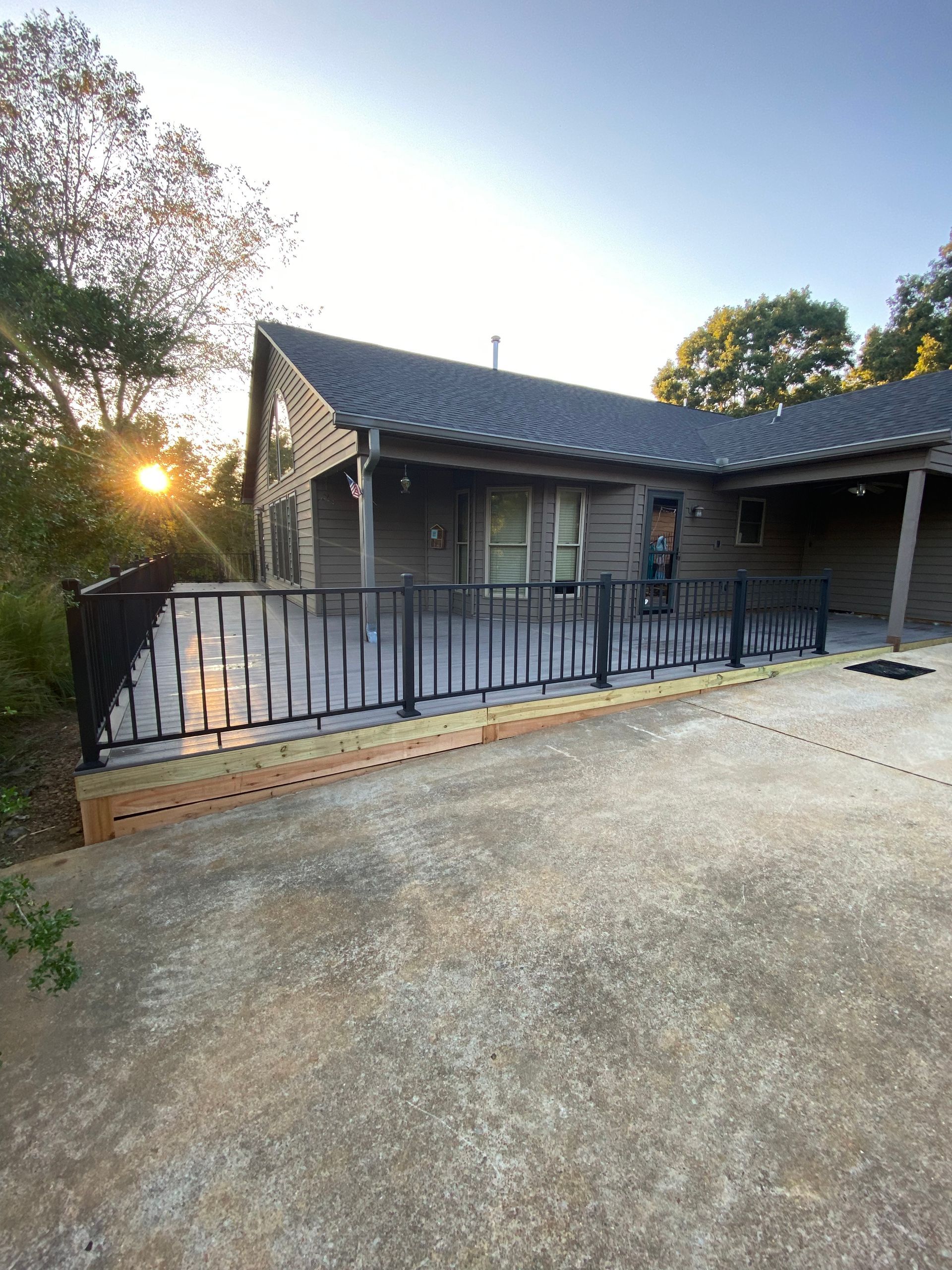 A cabin with a black metal railing, a deck, and a concrete driveway. The sun sets in the background.