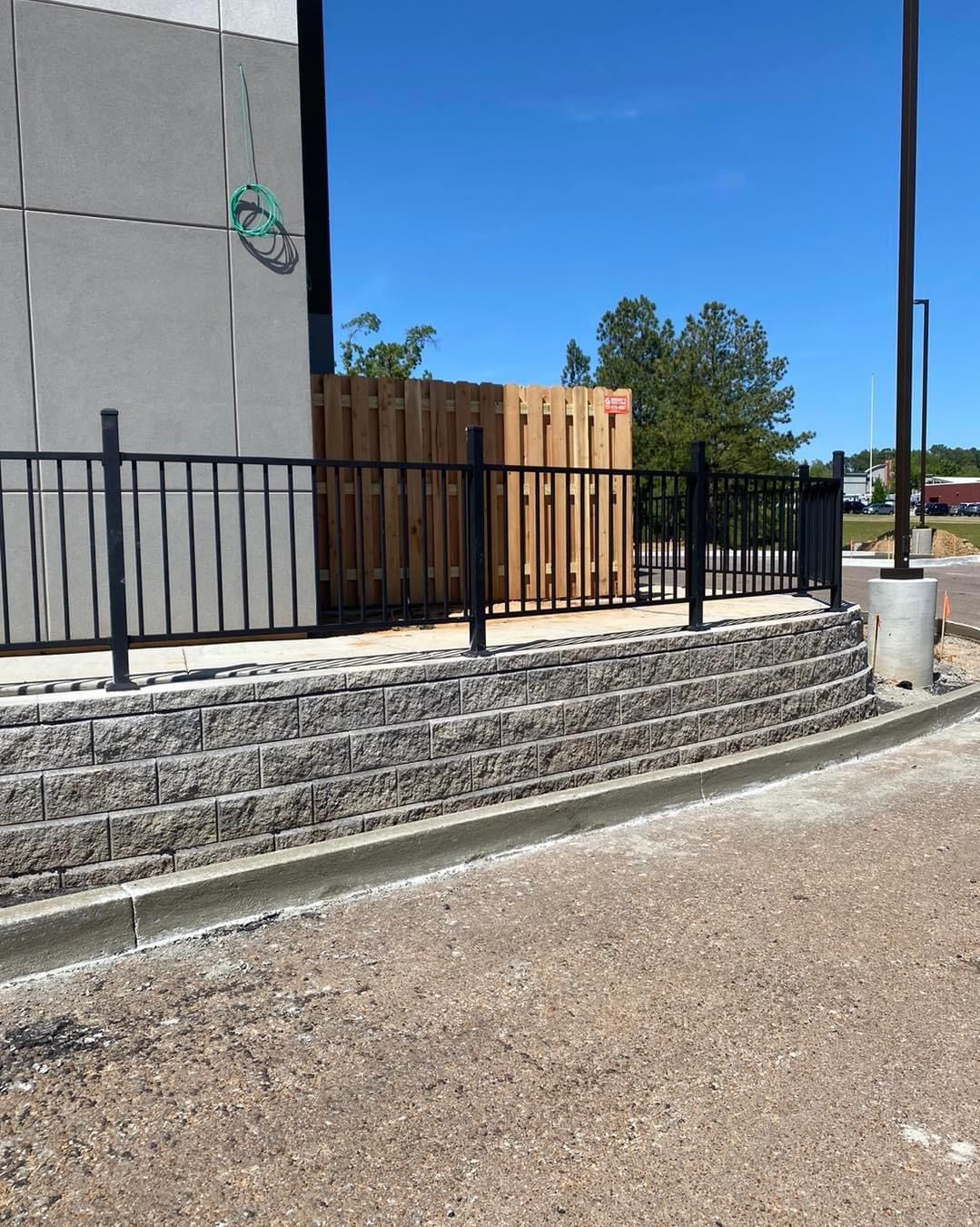 Black metal fence atop a retaining wall with wood fence and building in the background, under a blue sky.