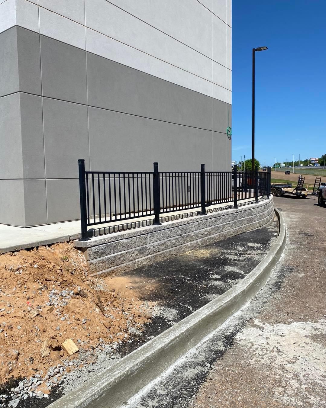 Gray building with black fence on retaining wall along concrete curb. Sunny day.
