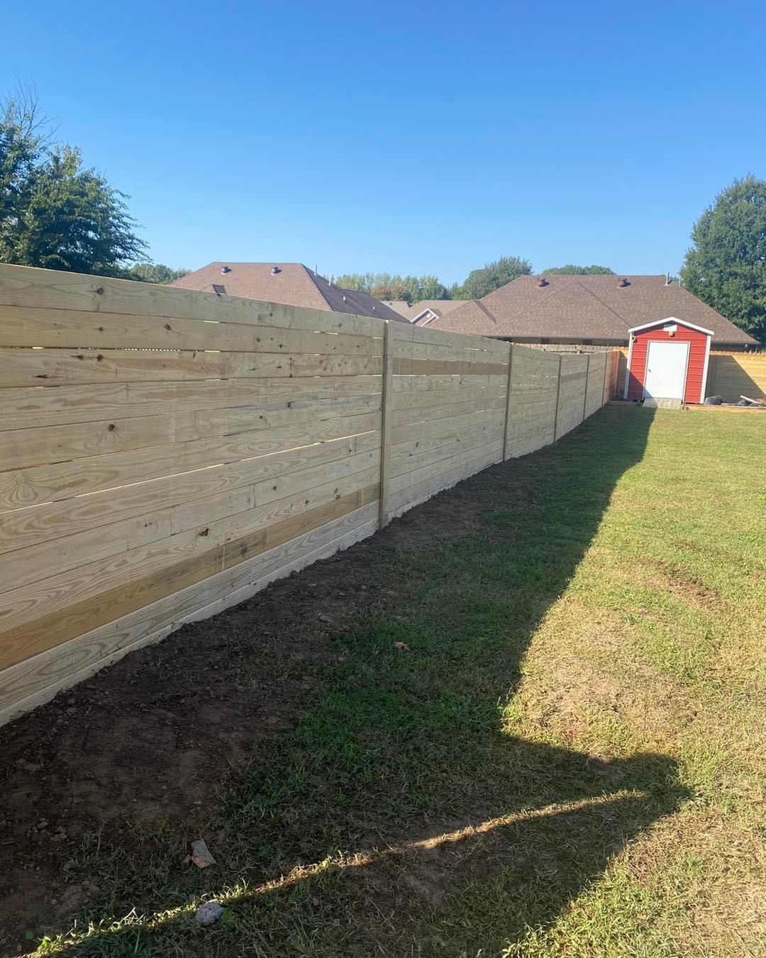 A wooden fence in a backyard on a sunny day with homes and a shed in the background.