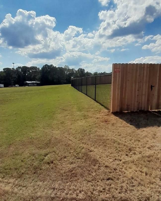 A chain-link fence alongside a wooden fence bordering a grassy field under a partly cloudy sky.