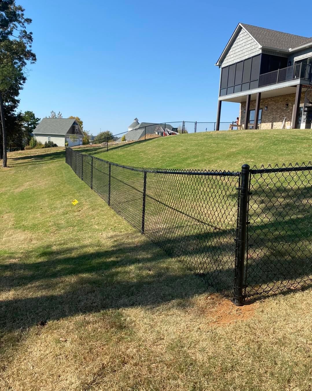 Black chain-link fence on a grassy hill, beside a house with a porch, under a clear blue sky.