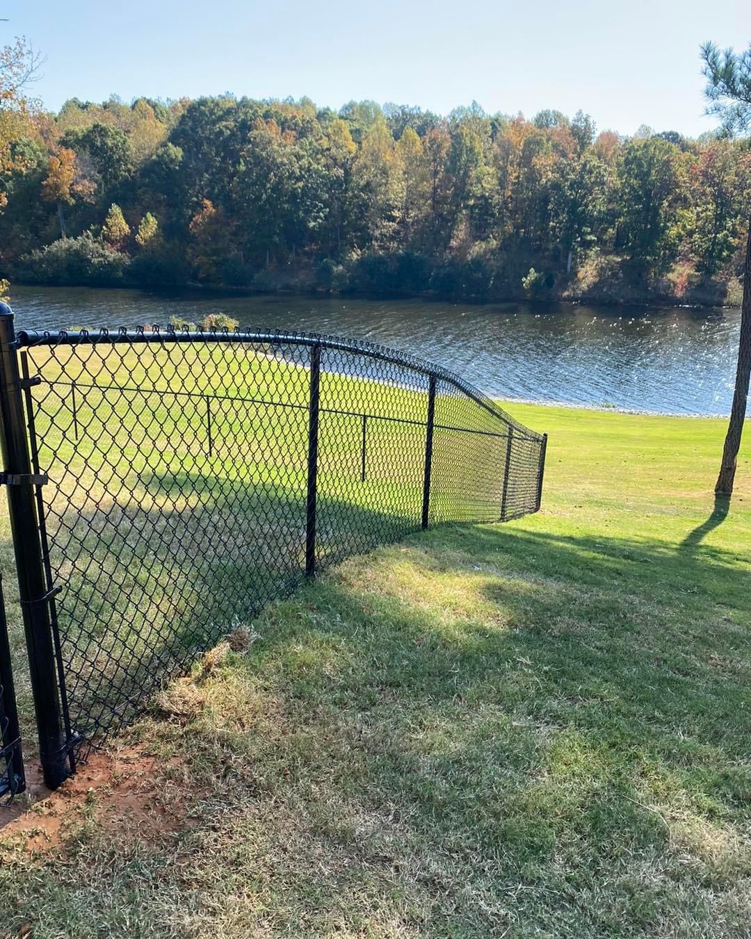 Black chain-link fence bordering a green grassy area next to a lake. Trees in the background.