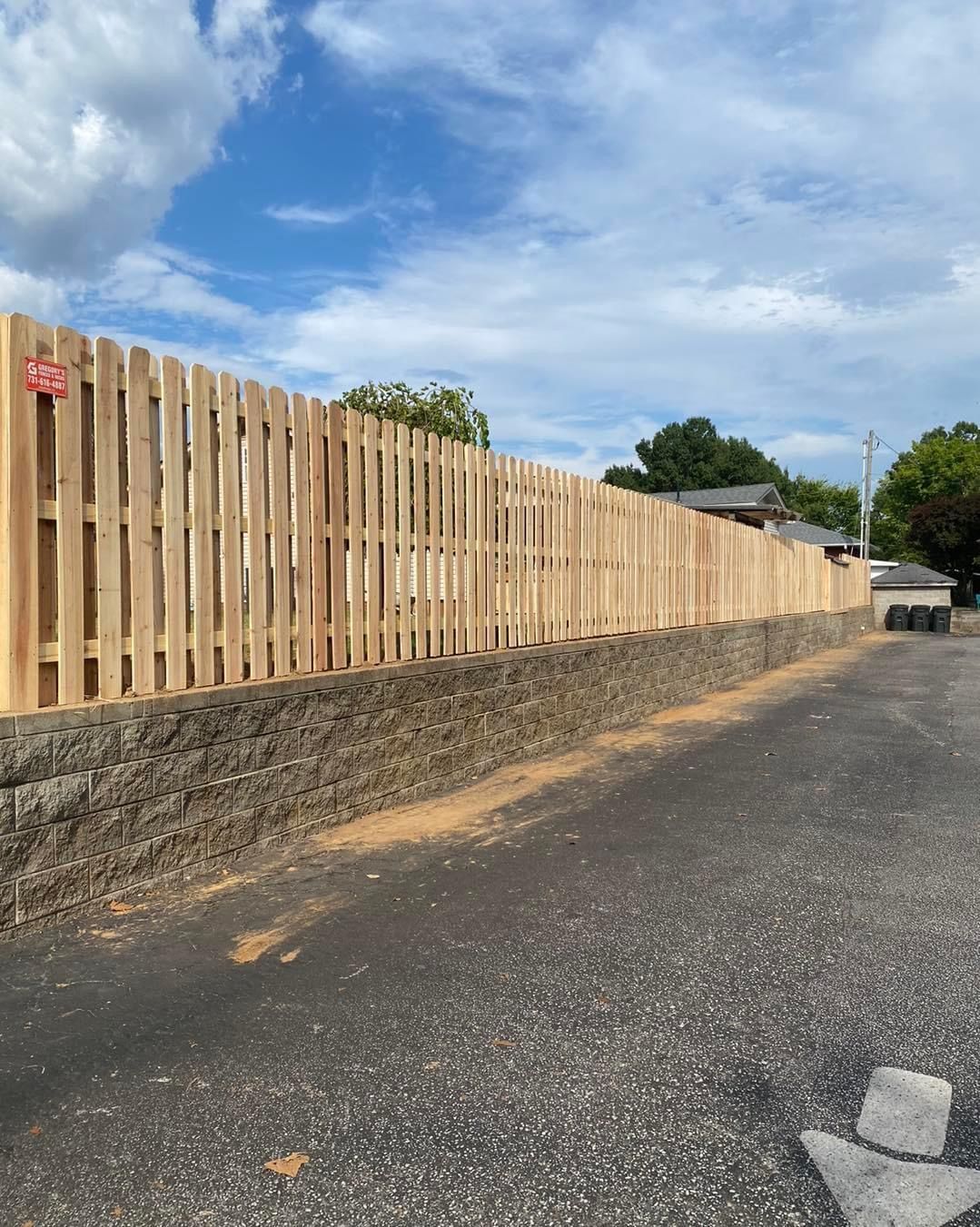 Wooden fence atop a retaining wall along a parking lot on a sunny day.