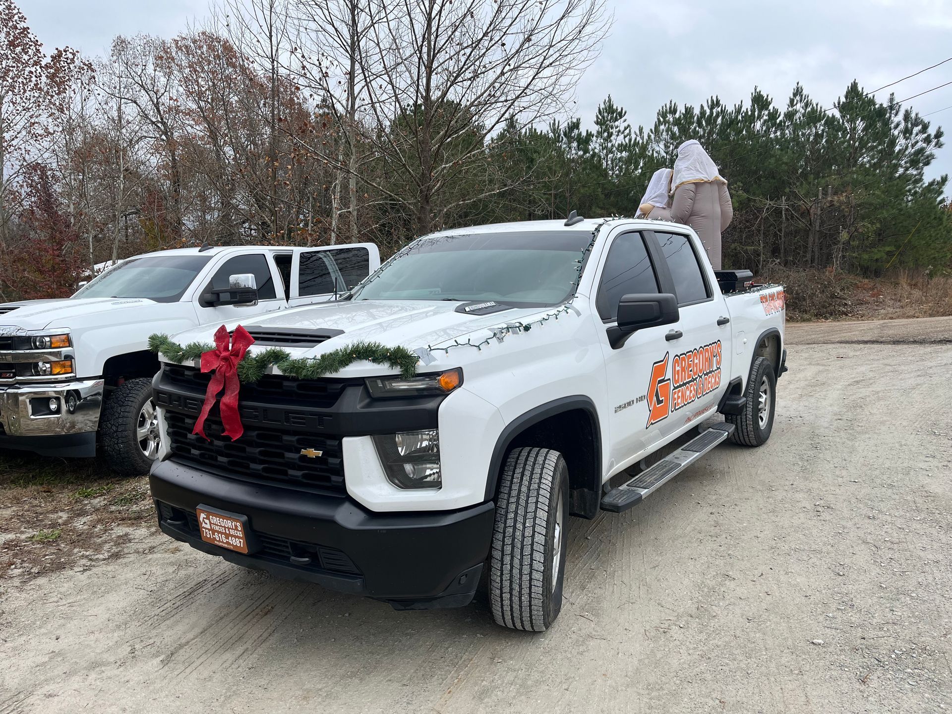 White work truck with a Christmas wreath, parked next to another truck on a dirt road.