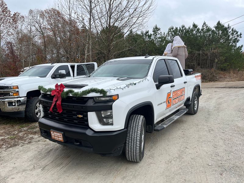 White Chevy truck decorated with a red bow and evergreen garland. Another truck is behind it. People in the bed.