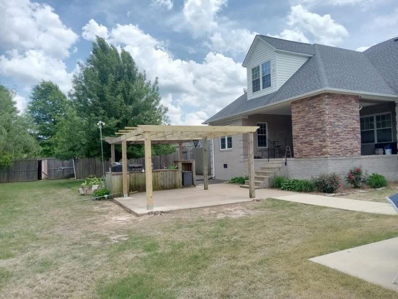 Pergola over a concrete patio in a backyard with a house, trees, and lawn. Cloudy sky.